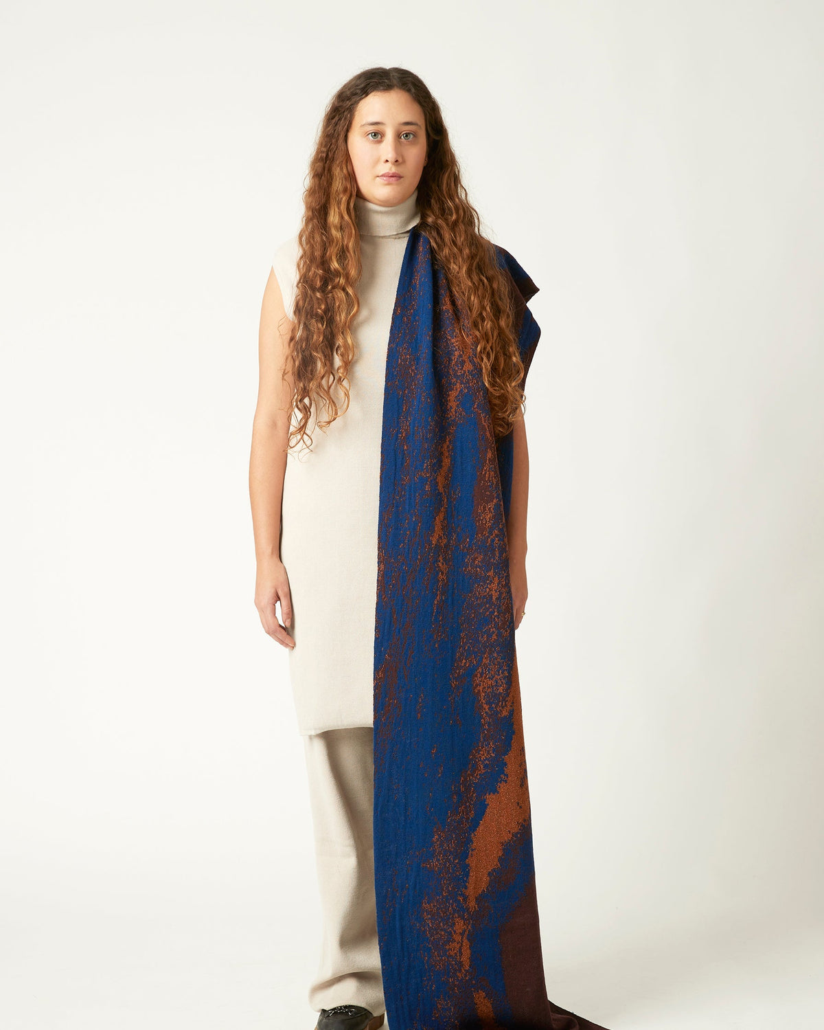 Wide shot of a young woman with a scarf draped over her shoulder. She's wearing a neutral light beige outfit and black shoes. The knit scarf is blue and brown with details in copper and gold.