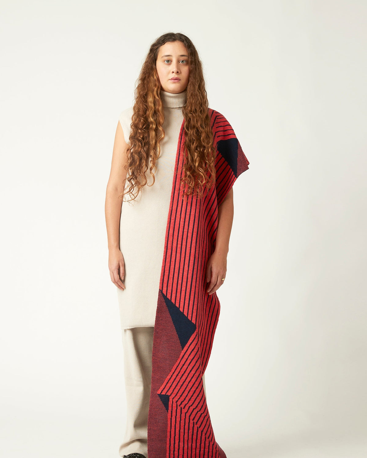 Wide shot of a young woman with a striped scarf draped over her shoulder. She's wearing a neutral light beige outfit and black shoes. The striped knitted scarf is coral pink and dark blue.