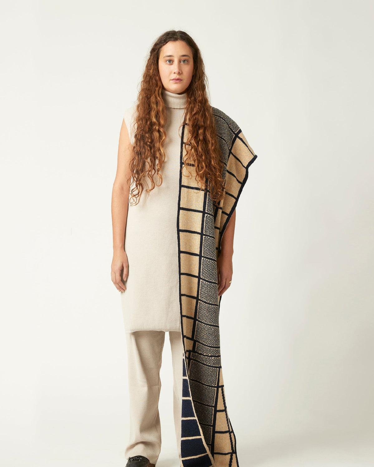 Wide shot of a young woman with a knitted scarf draped over her shoulder. She's wearing a neutral light beige outfit and black shoes. The knit scarf has an abstract pattern in dark blue and peach.