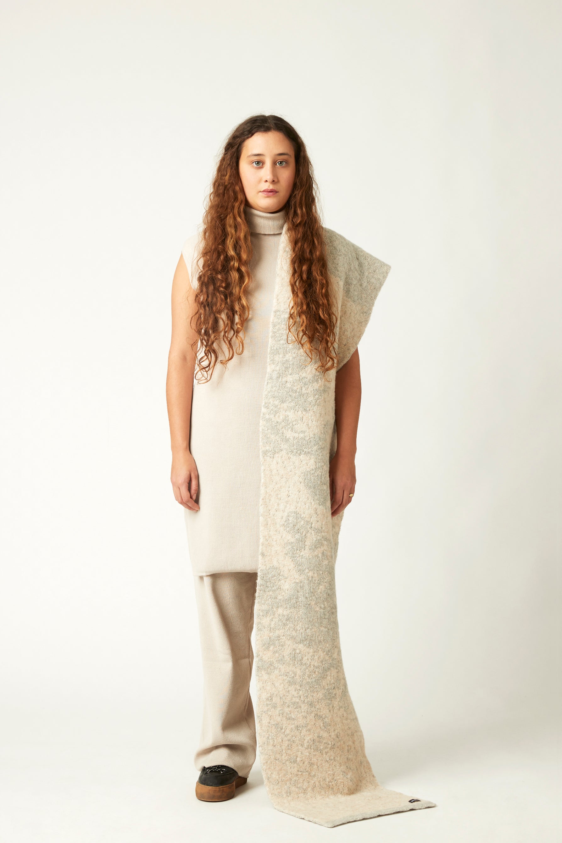 Full body shot of a young woman with a knitted scarf draped over the shoulder. She's wearing a neutral toned outfit and black shoes. The white and grey knit scarf is fluffy and bulky.