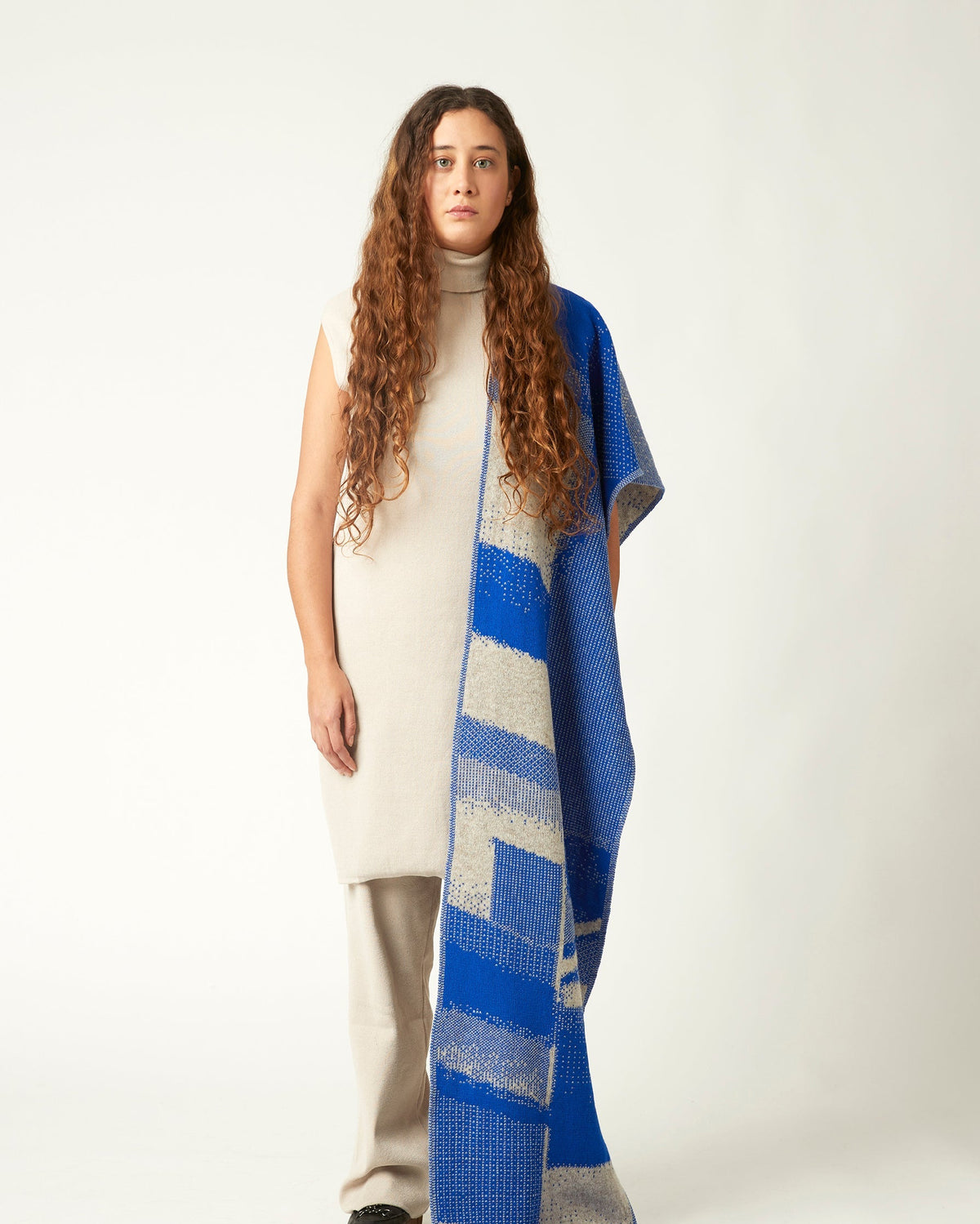 Wide shot of a young woman with a knit scarf draped over the shoulder. She's wearing a neutral toned outfit and black shoes. Packhshot of a jacquard knit scarf in cobalt blue and beige. The scarf has an abstract geometric bitmap pattern.