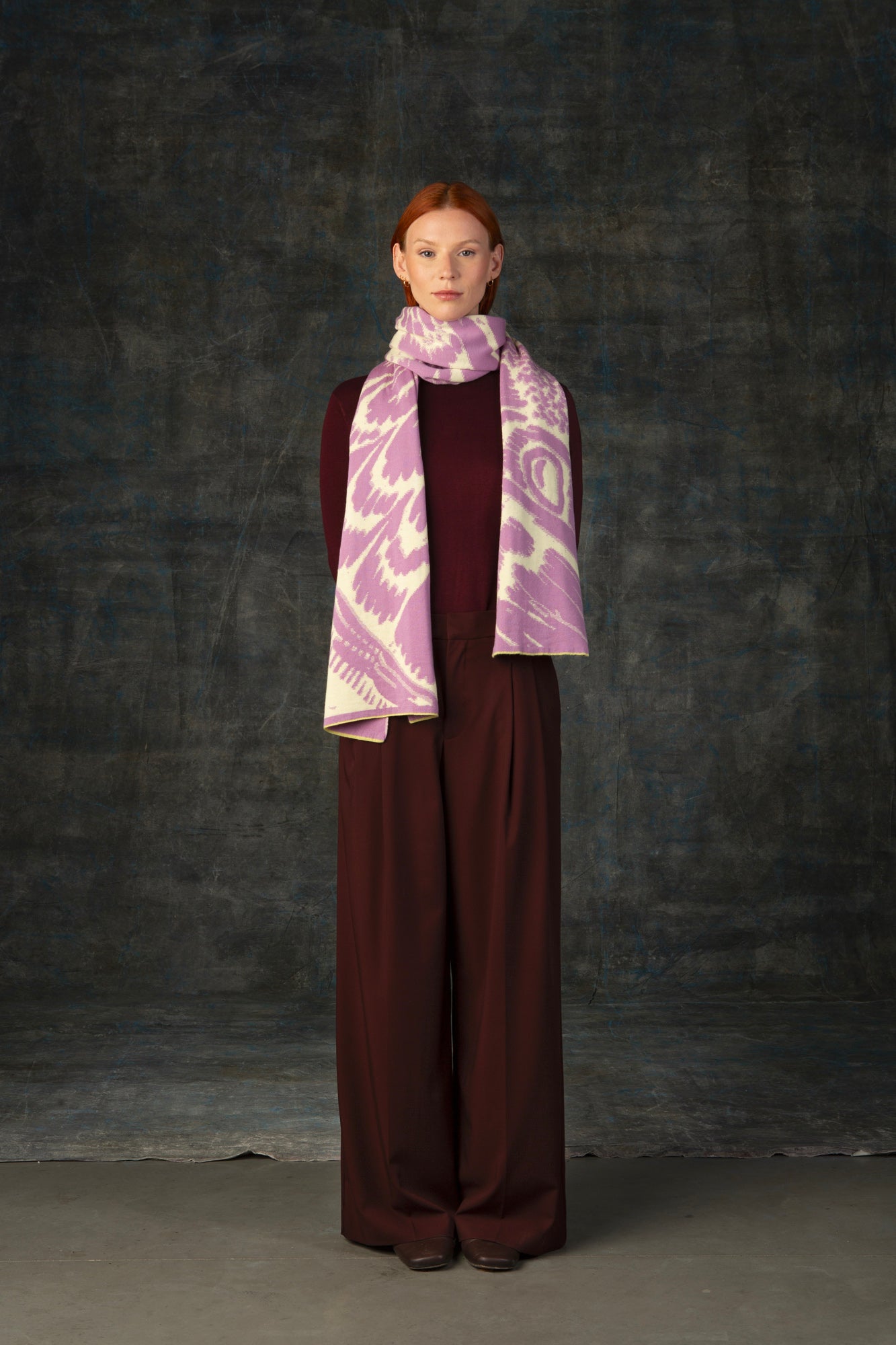 Female model wearing a burgundy outfit with a pink and beige maxi scarf, with an organic pattern, draped around her neck.