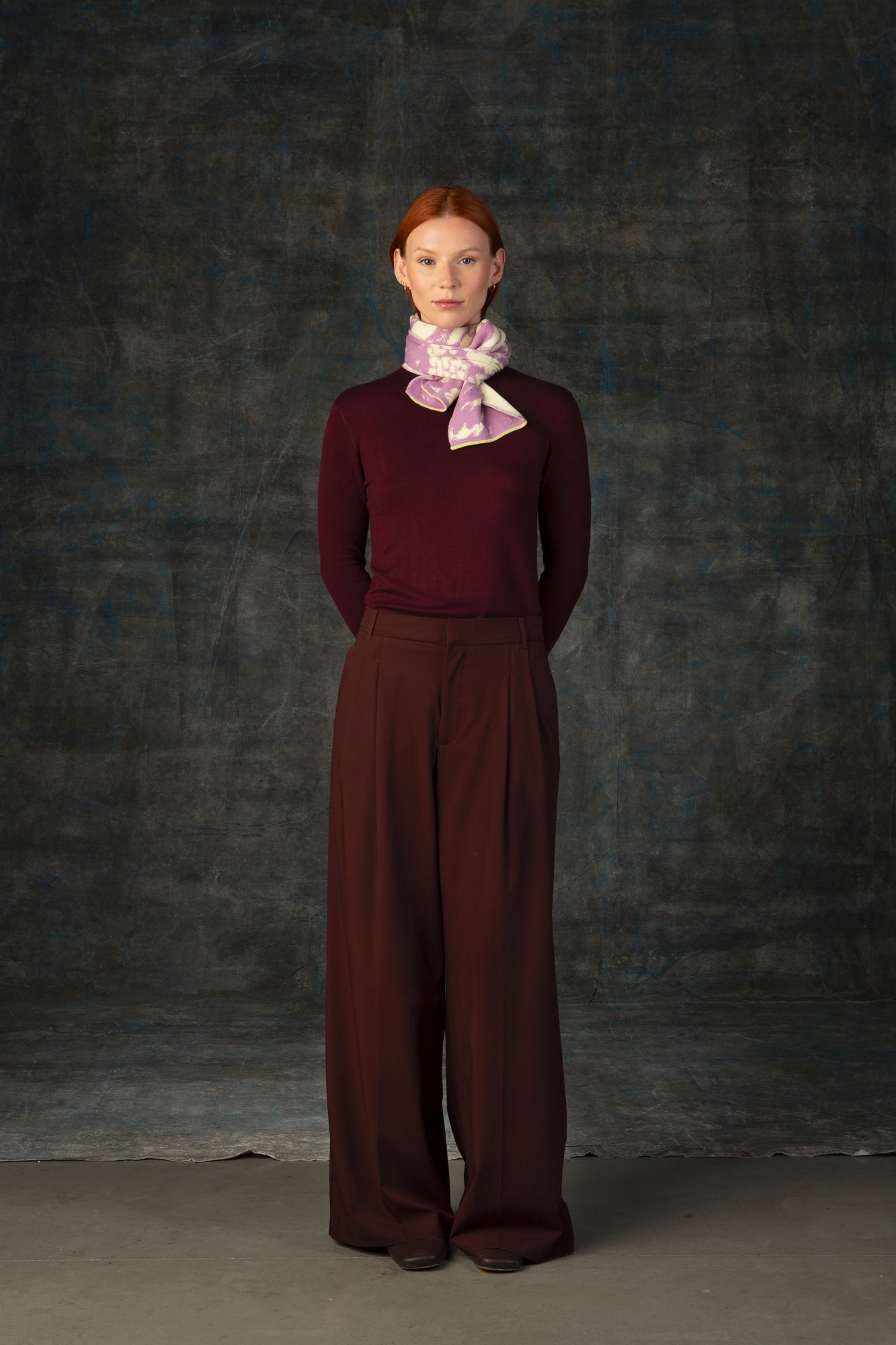 Female model wearing a burgundy outfit with a pink and beige mini scarf, with an organic pattern, tied around her neck.