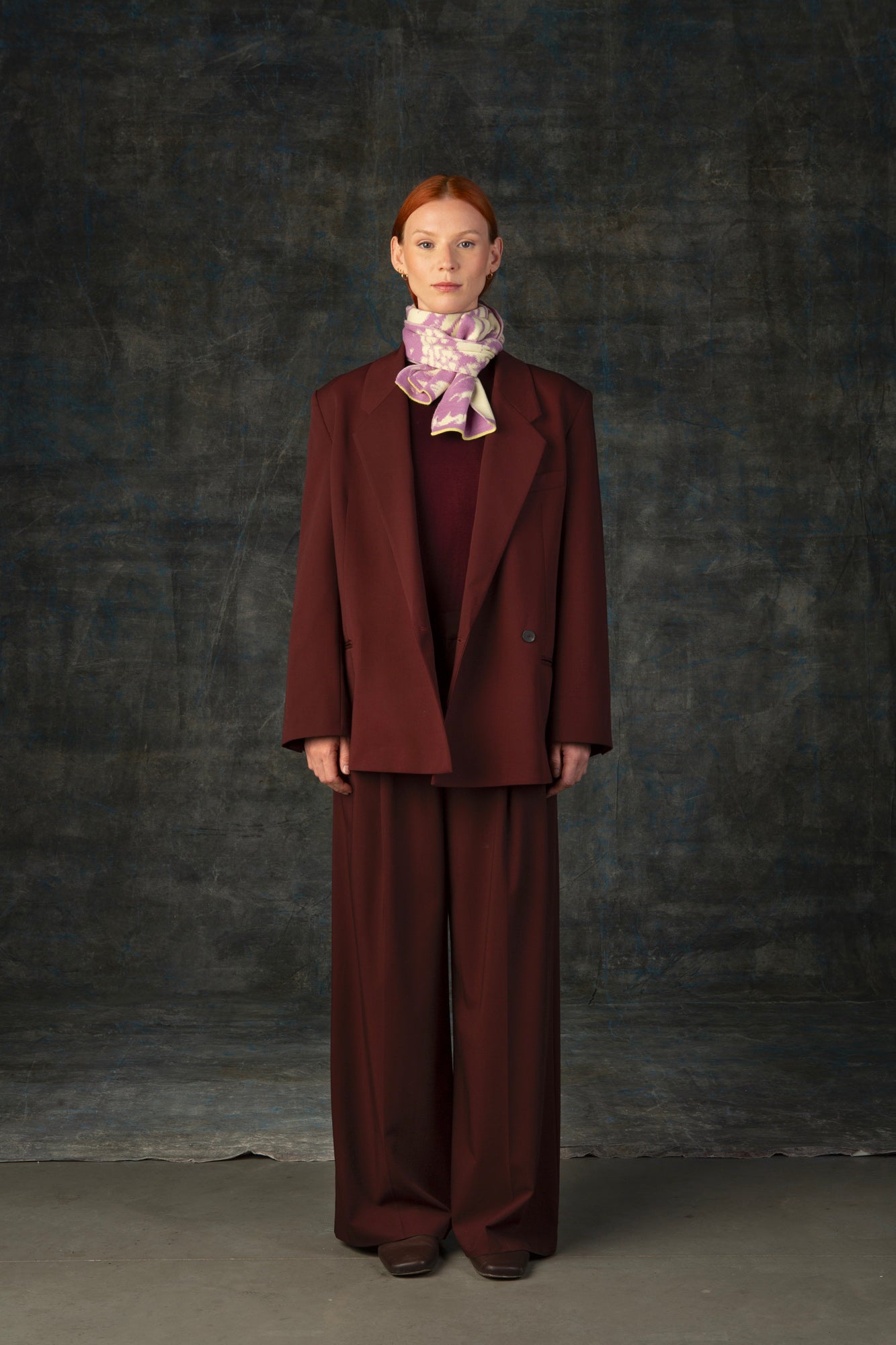 Female model wearing a rust outfit with a pink and beige mini scarf, with an organic pattern, tied around her neck.