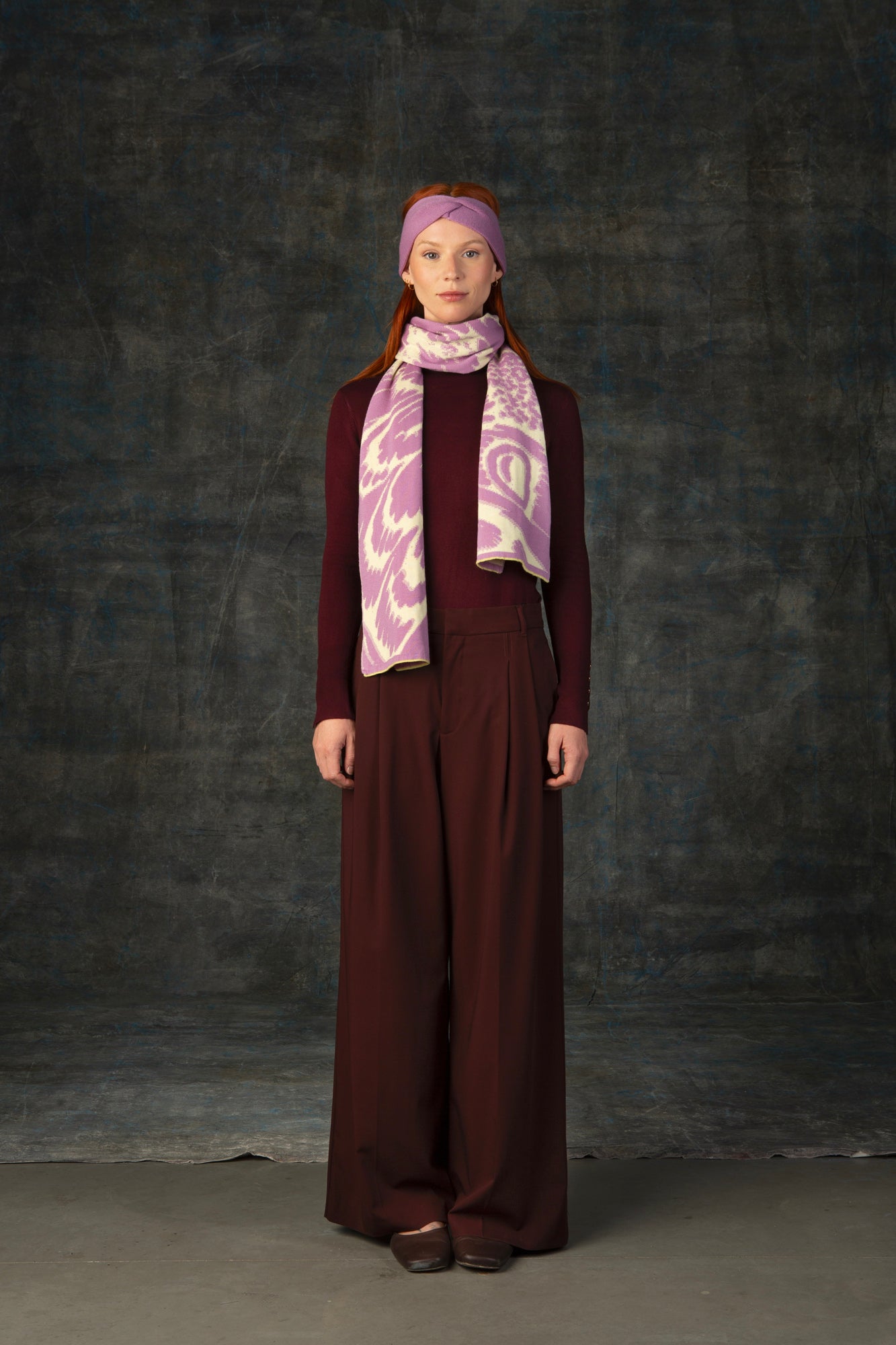 Female model wearing a burgundy outfit with a mauve pink and beige scarf with an organic pattern, draped around her neck and matching pink headband.