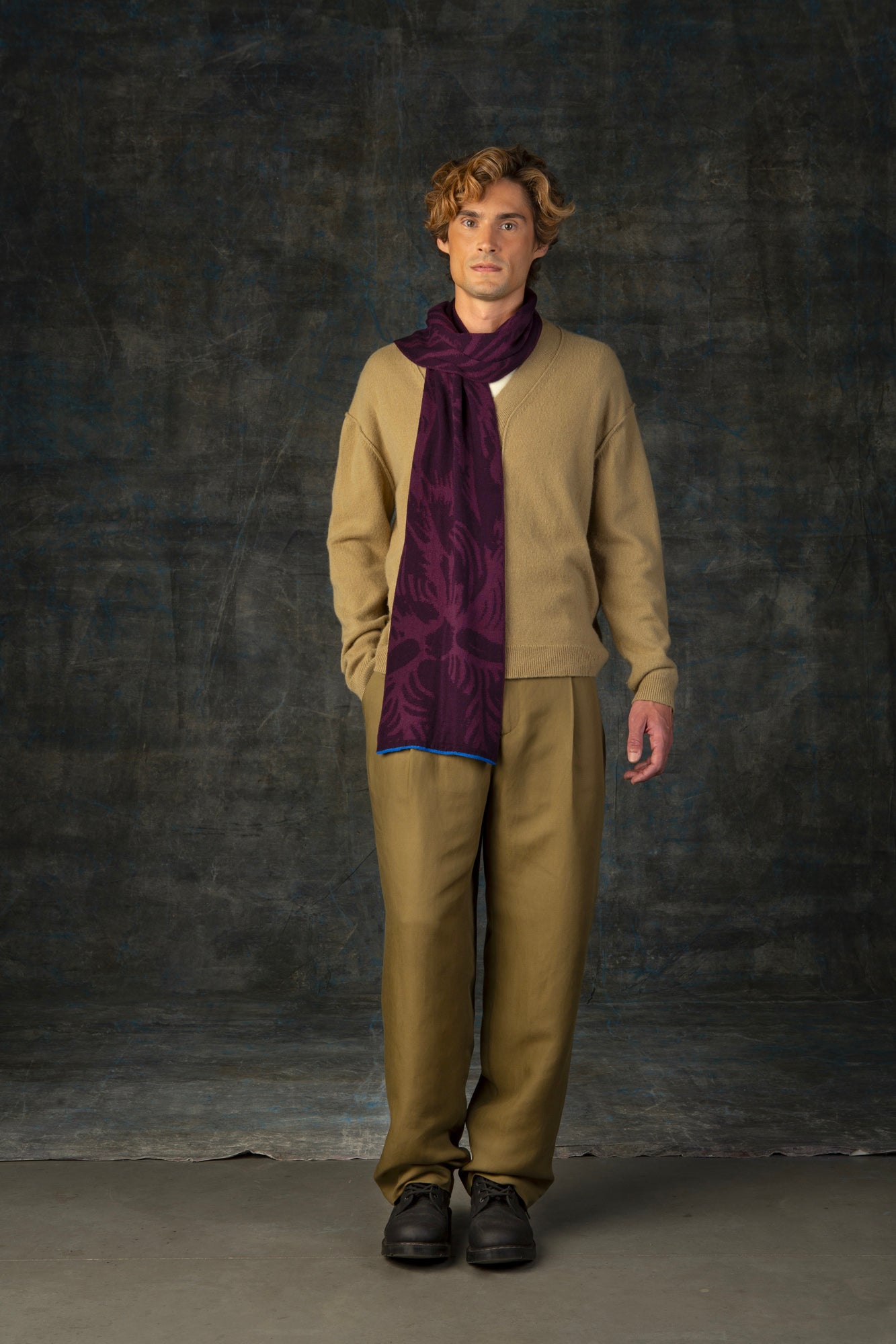 Male model wearing a muted mustard yellow outfit with a dark purple midi scarf, with nature-inspired pattern draped around over his shoulder.