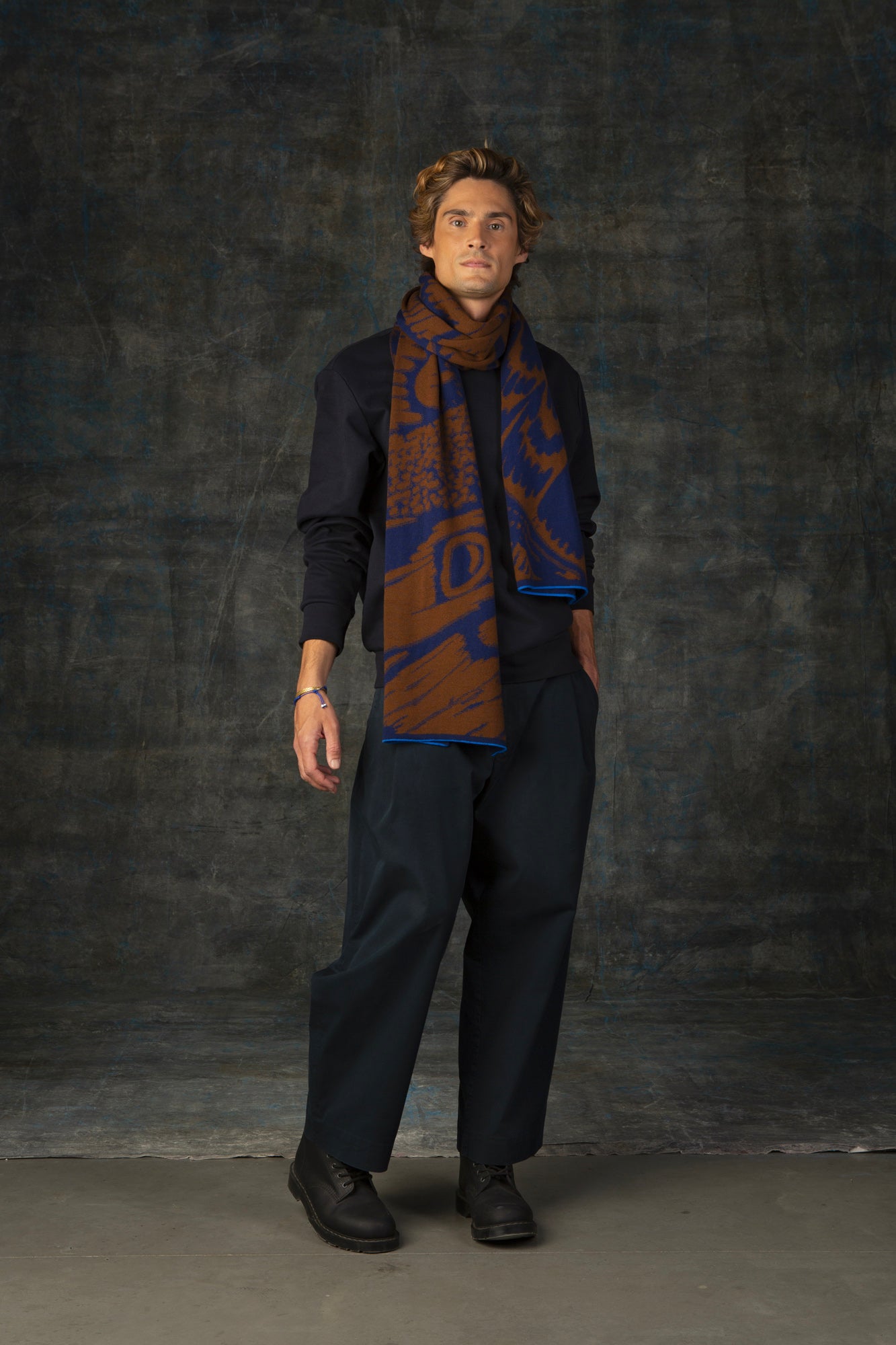 Male model wearing a dark blue outfit with a brown and blue maxi scarf, with an organic pattern, draped around his neck.