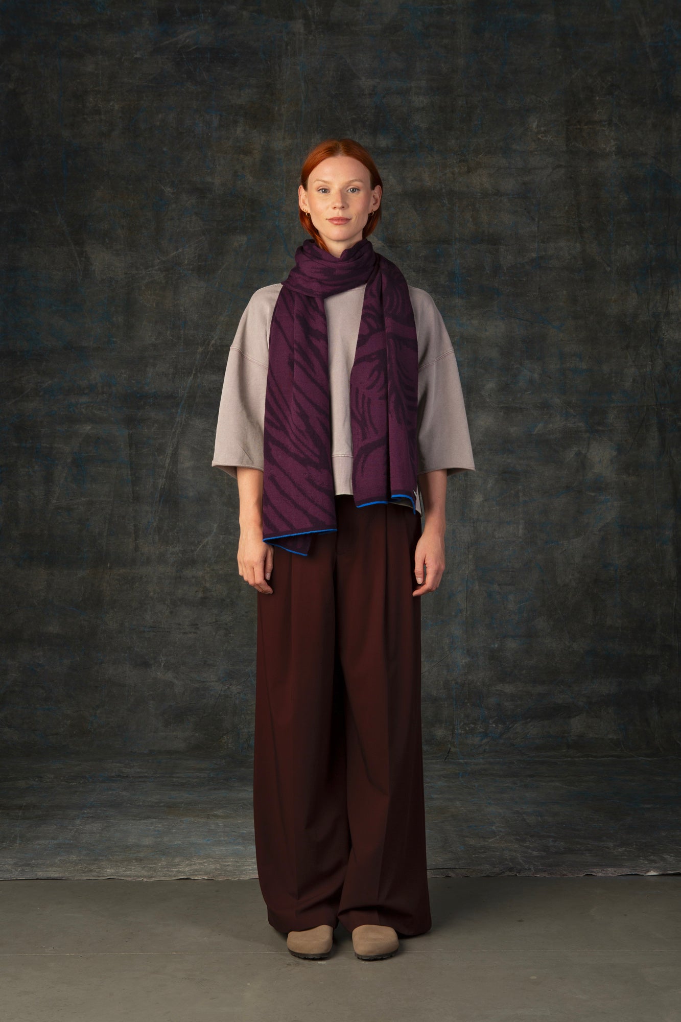 Female model wearing a neutral outfit with a dark purple maxi scarf, with nature-inspired pattern draped around her neck, against a dark background.