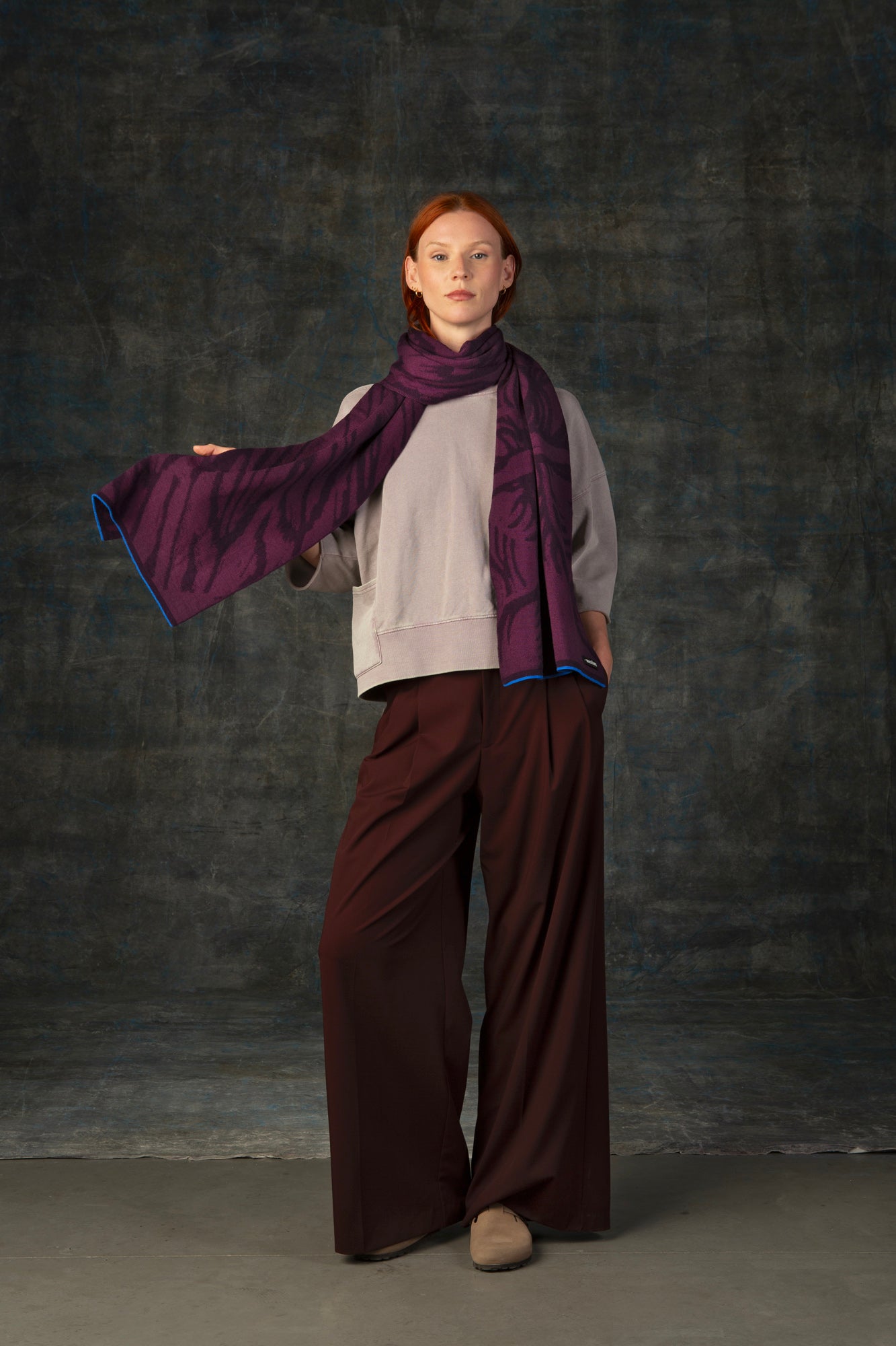 Female model wearing a neutral outfit with a dark purple maxi scarf, with nature-inspired pattern draped around her neck, against a dark background.