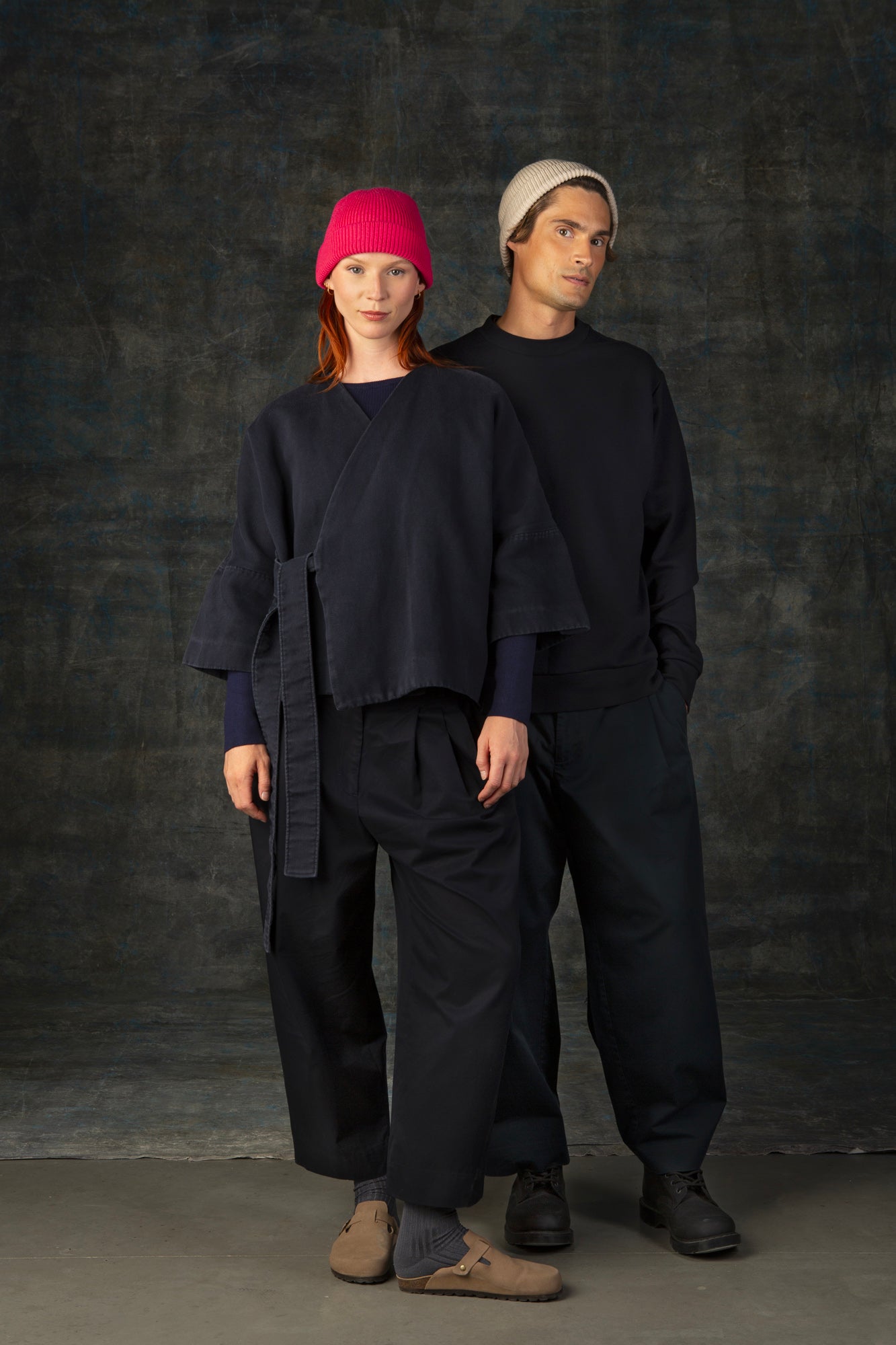 Two people wearing dark clothing and beanies against a dark background. The female model is wearing a bright pink beanie. The male model is wearing an ecru beanie.