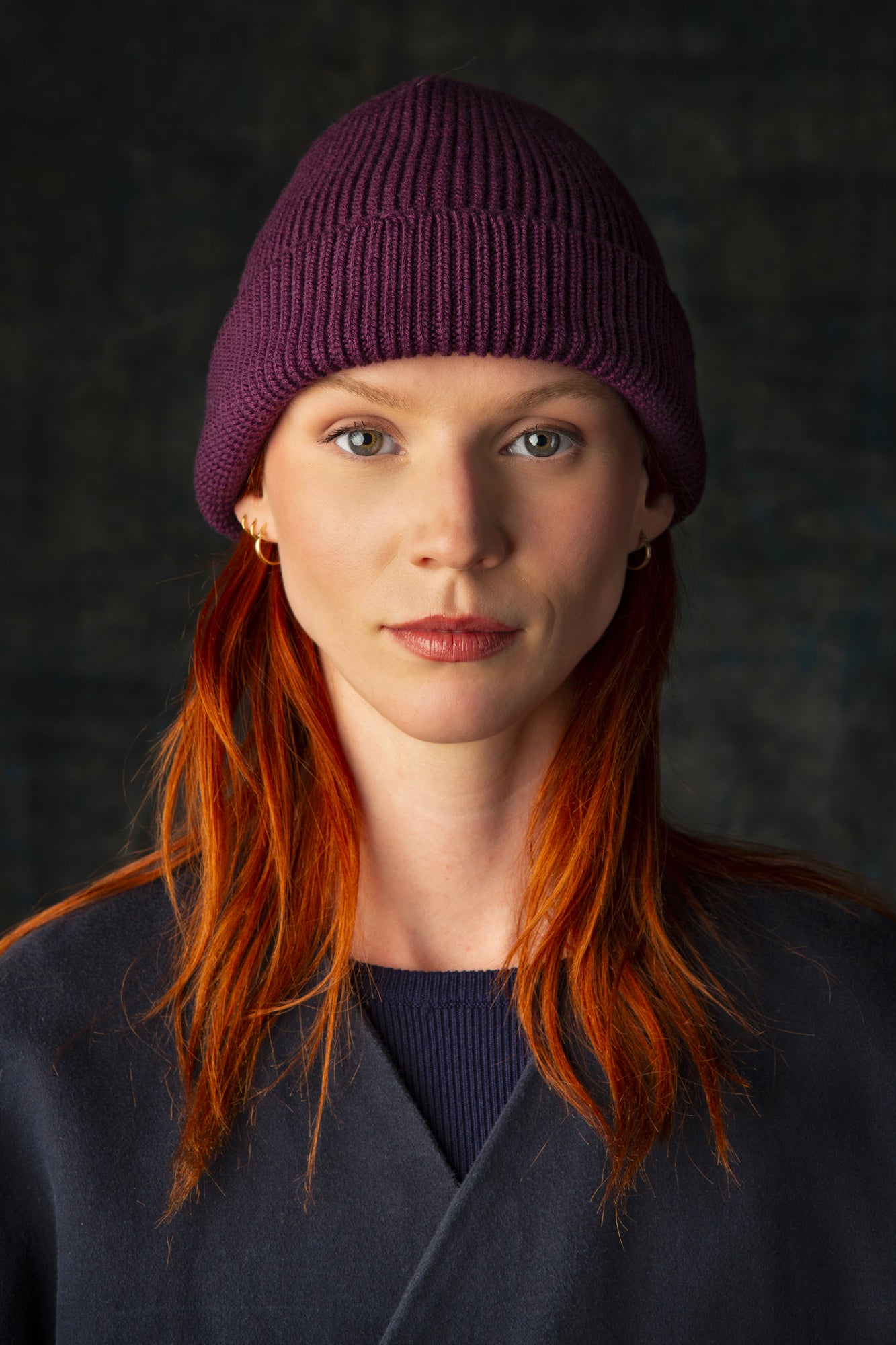 Person wearing a grape purple knit beanie and dark blue coat against a dark background.