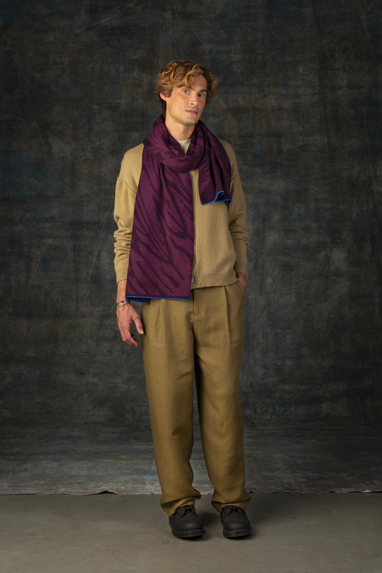 Male model wearing a muted mustard yellow outfit with a dark purple maxi scarf, with nature-inspired pattern draped around his neck, against a dark background.