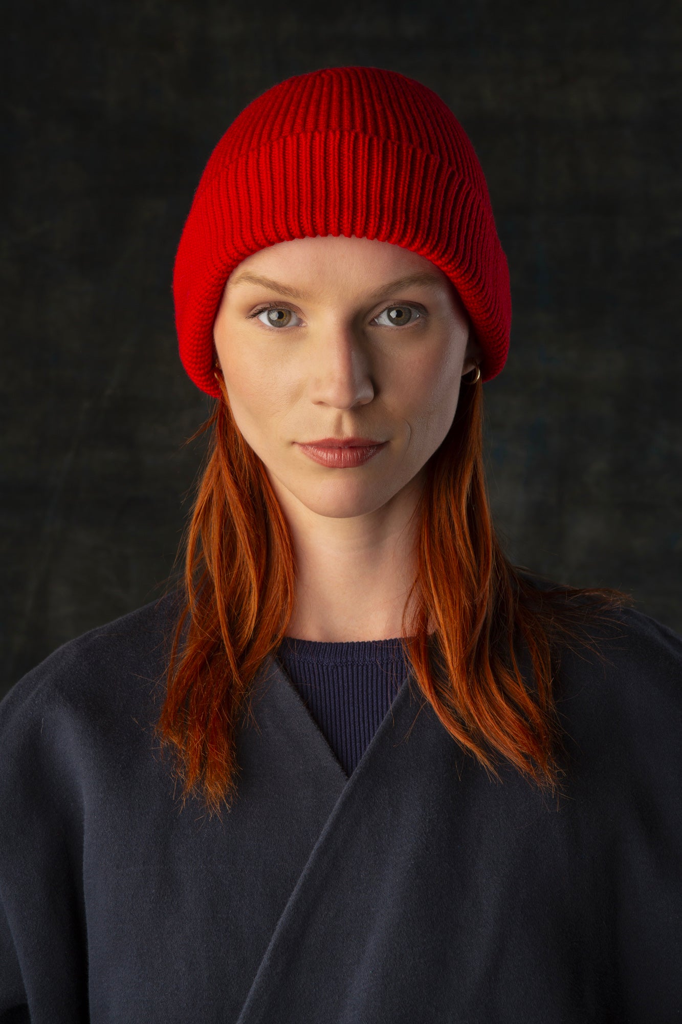 Person wearing a bright red knit beanie and dark blue coat against a dark background.
