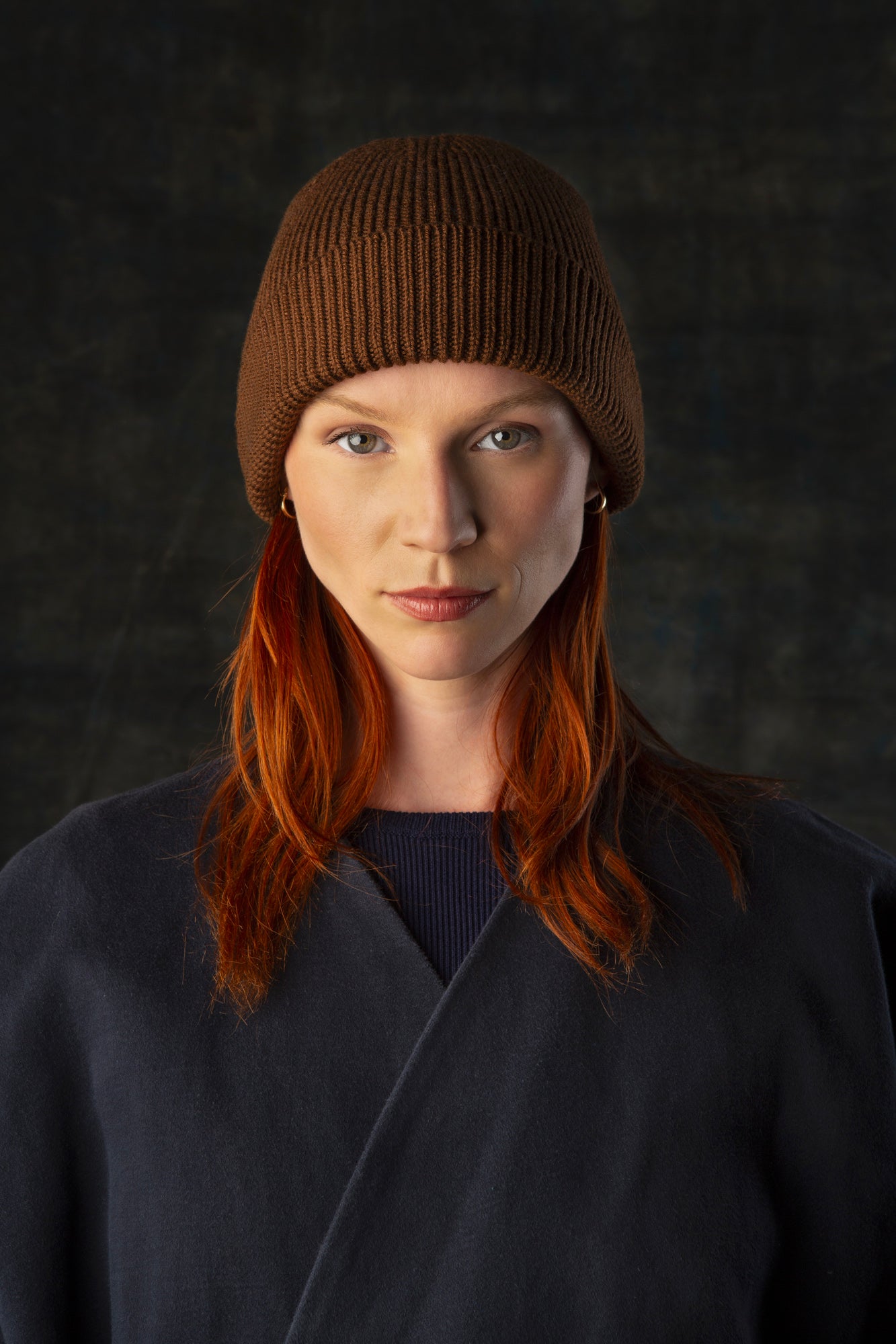Person wearing a cacao brown knit beanie and dark blue coat against a dark background.