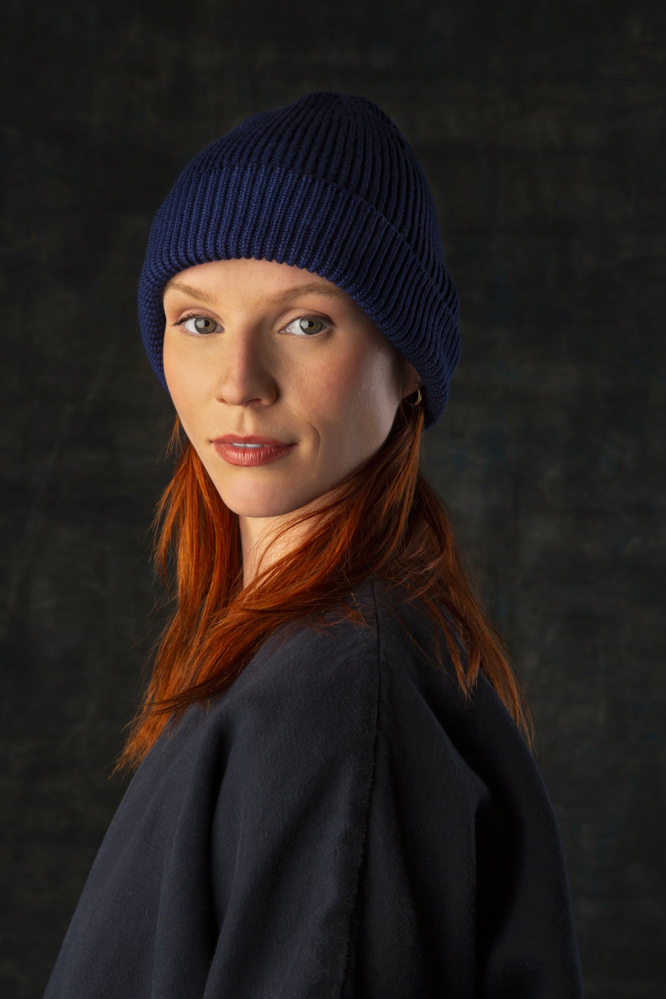 Person wearing a dark indigo blue knit beanie and dark blue coat against a dark background.