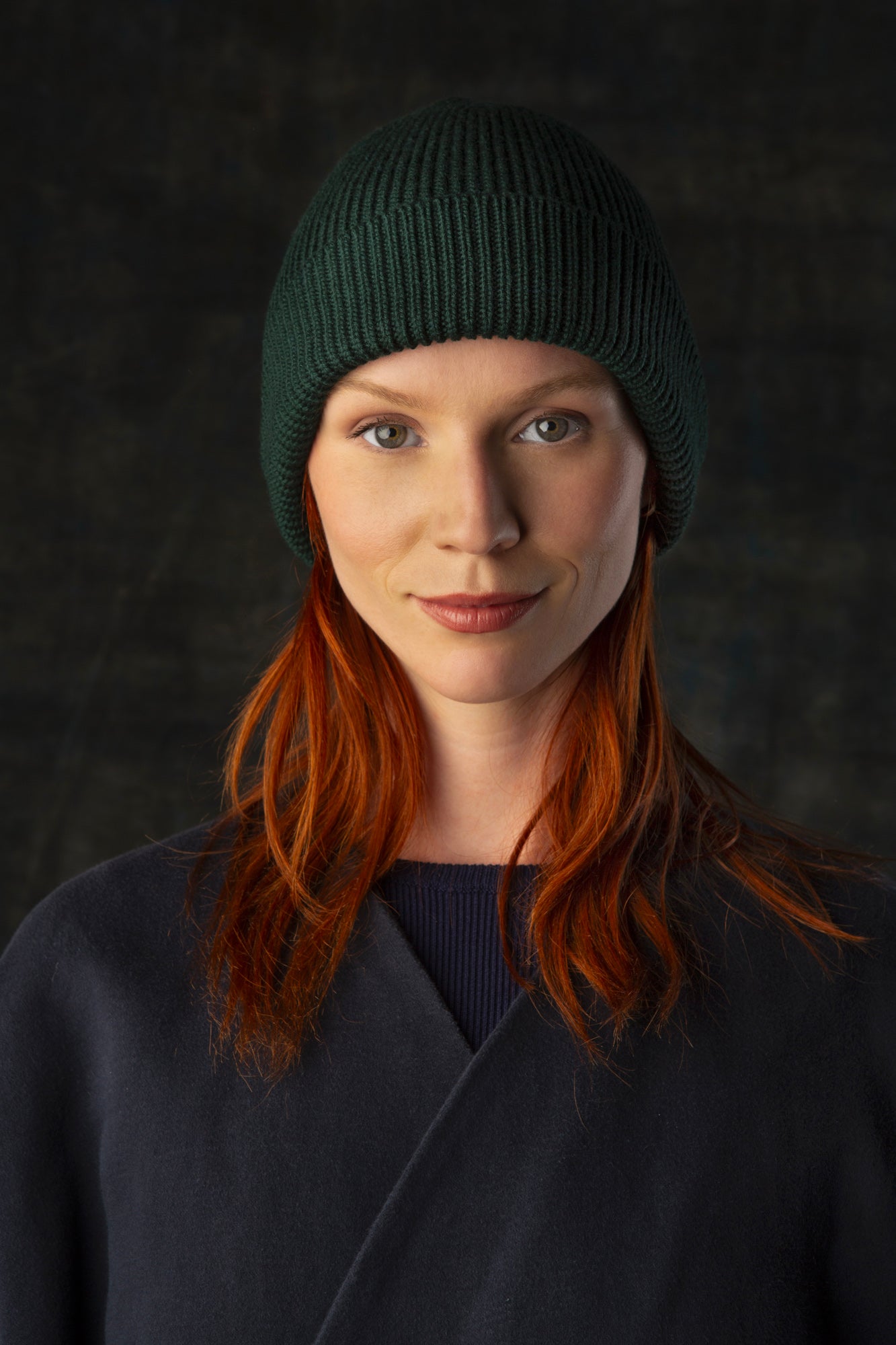 Person wearing a dark forest green knit beanie and dark blue coat against a dark background.