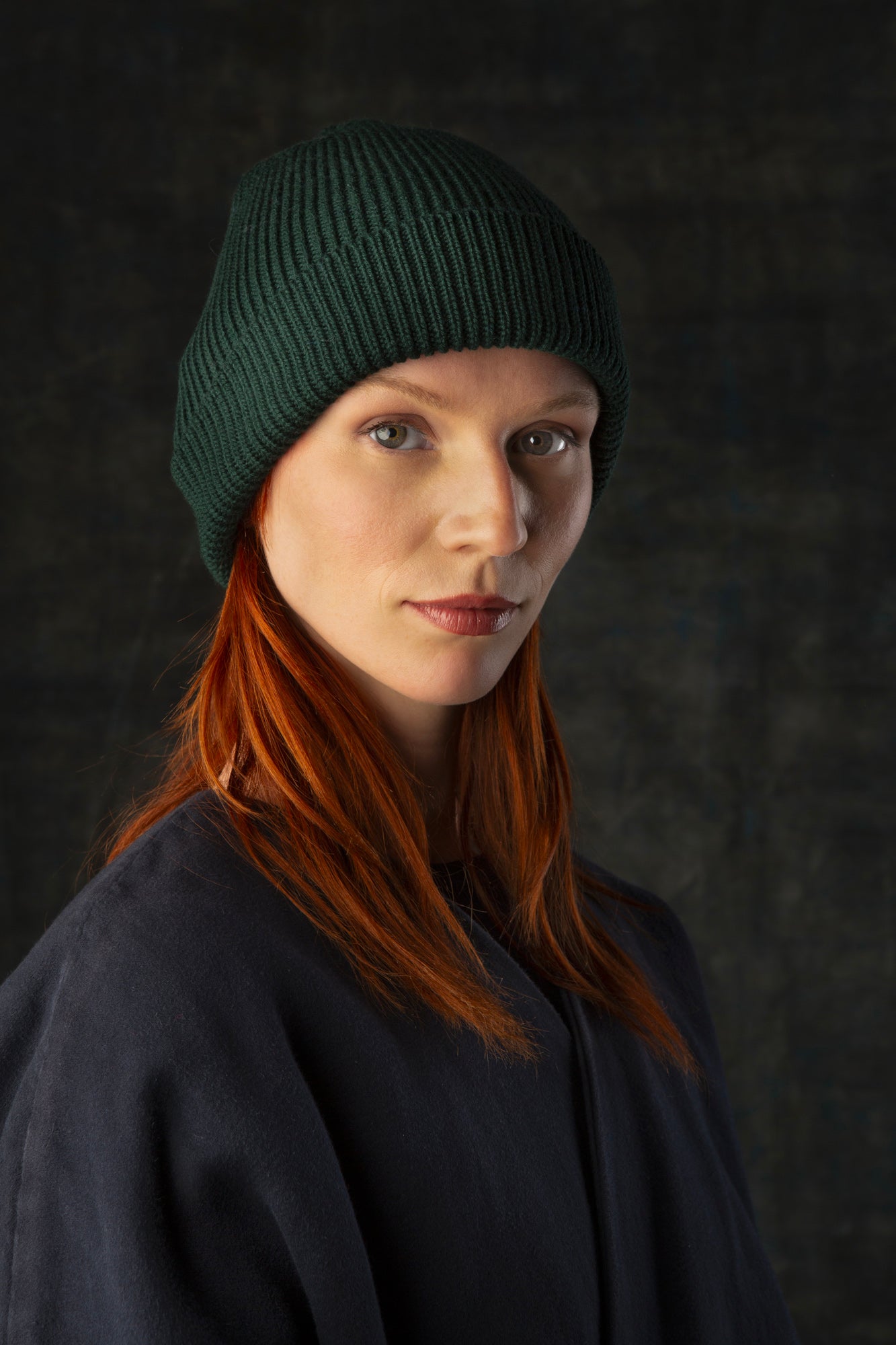 Person wearing a forest green knit beanie and dark blue coat against a dark background.