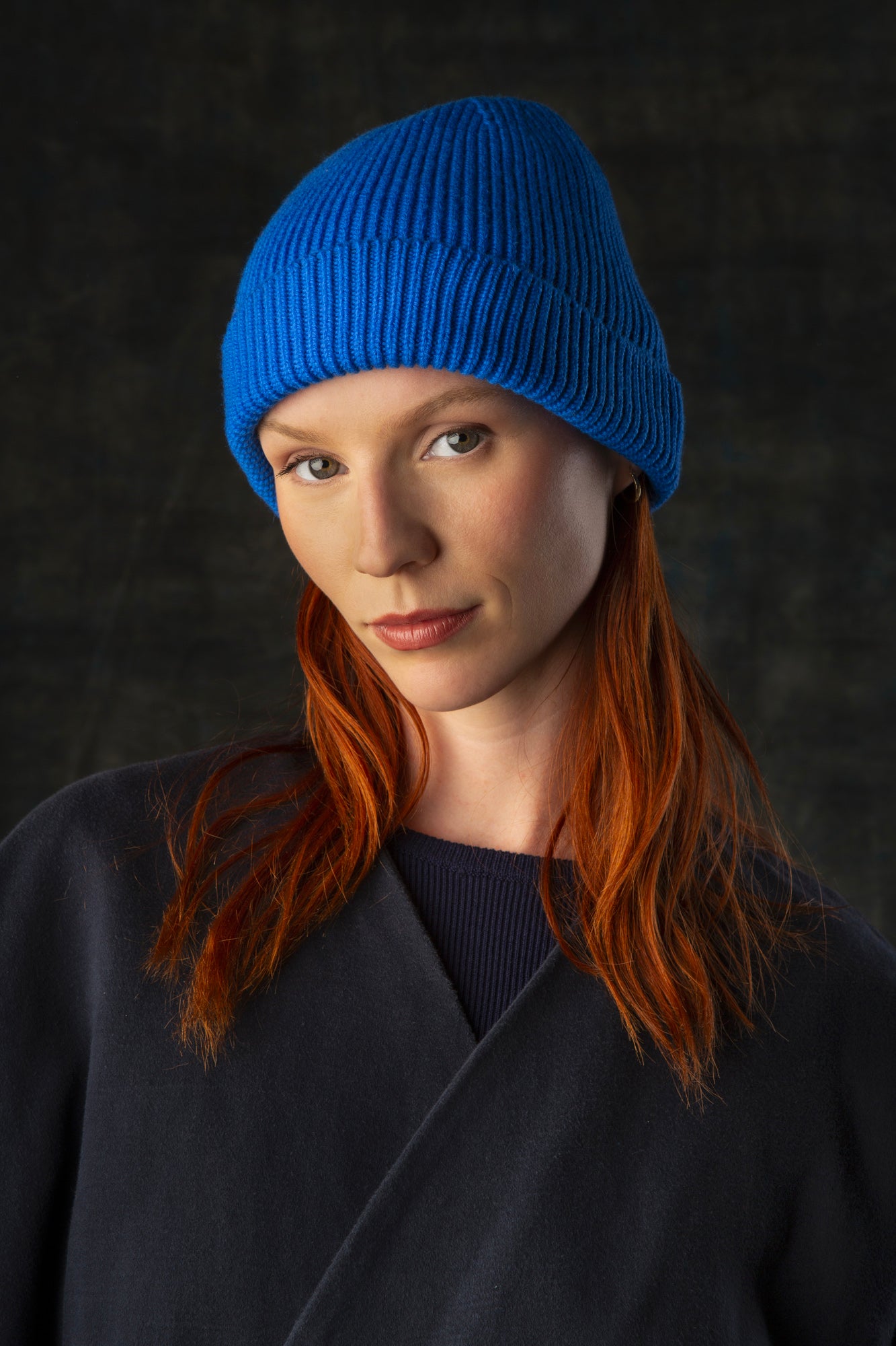 Person wearing a cobalt blue knit beanie and dark blue coat against a dark background.