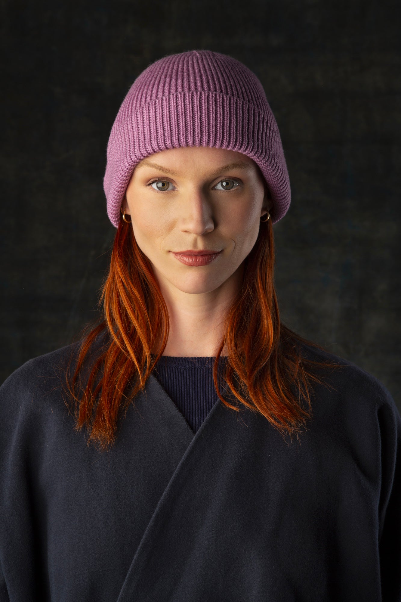 Person wearing a mauve pink knit beanie and dark blue coat against a dark background.