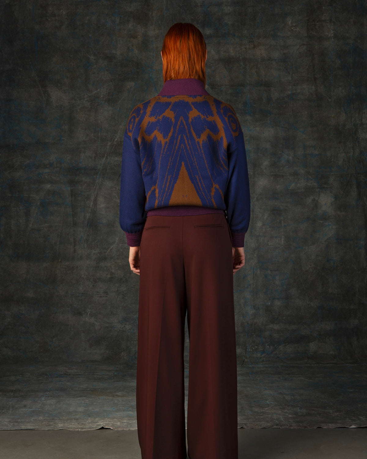 Female model facing away from the camera wearing a cacao brown bomber jacket with dark blue sleeves and purple cuffs. There is an organic pattern inspired by a butterfly's wings on the back of the jacket.