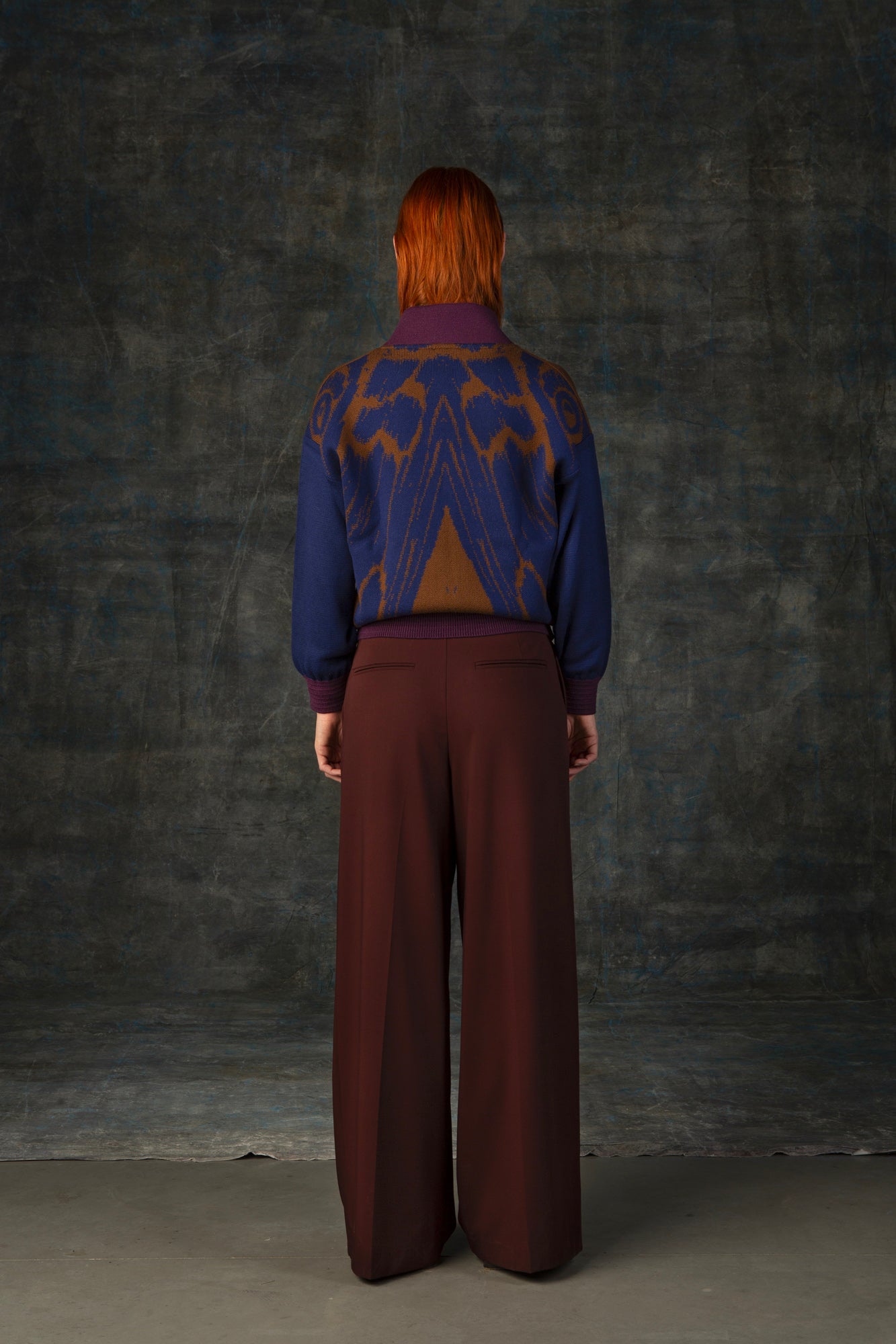 Female model facing away from the camera wearing a cacao brown bomber jacket with dark blue sleeves and purple cuffs. There is an organic pattern inspired by a butterfly's wings on the back of the jacket.