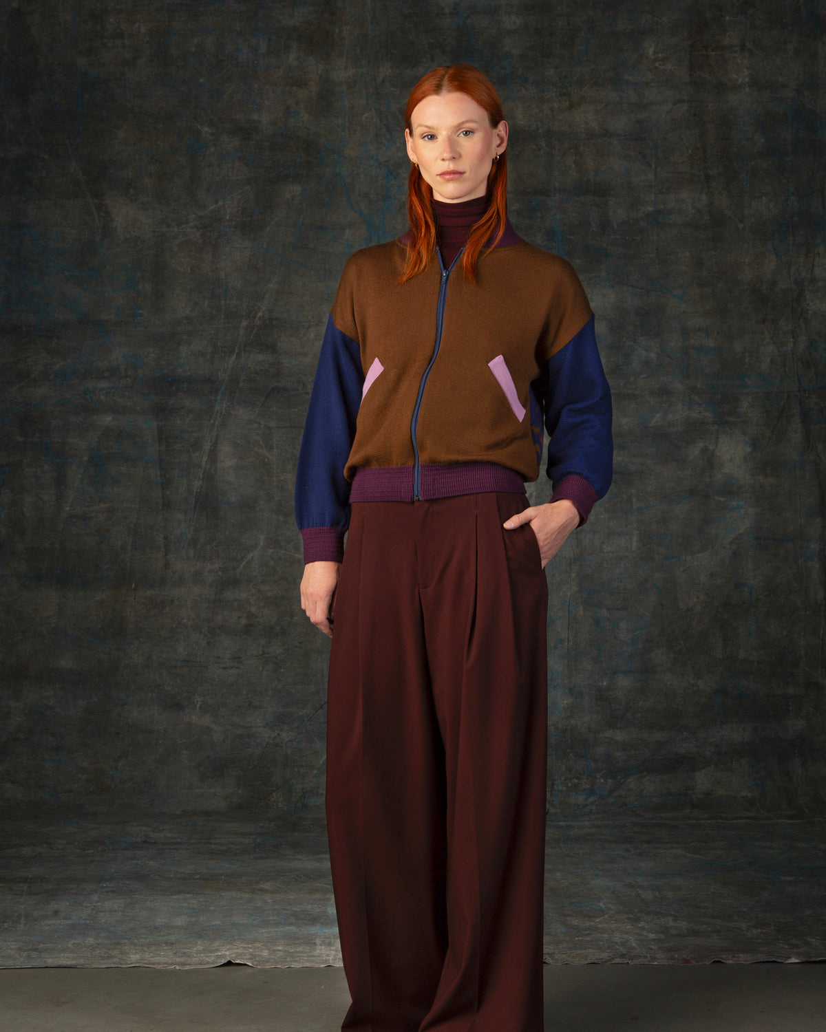 Female model wearing a cacao brown zip-up bomber jacket with dark blue sleeves and purple cuffs.