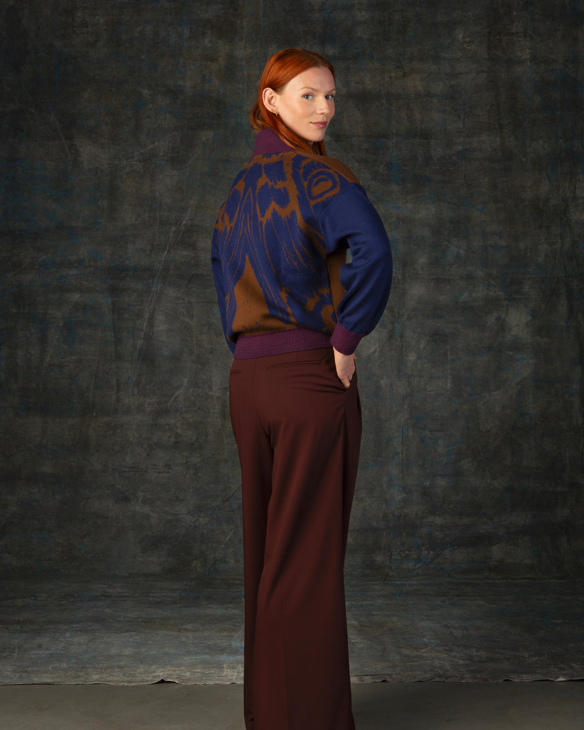 Female model wearing a cacao brown bomber jacket with dark blue sleeves and purple cuffs. There is an organic pattern inspired by a butterfly's wings on the back of the jacket.