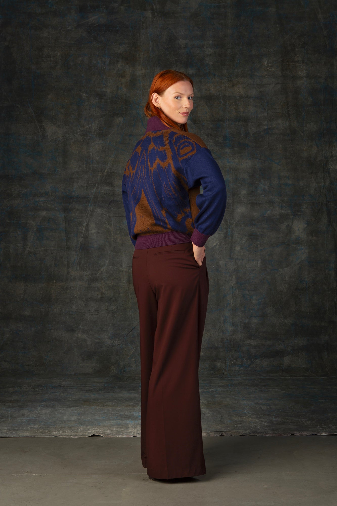 Female model wearing a cacao brown bomber jacket with dark blue sleeves and purple cuffs. There is an organic pattern inspired by a butterfly's wings on the back of the jacket.