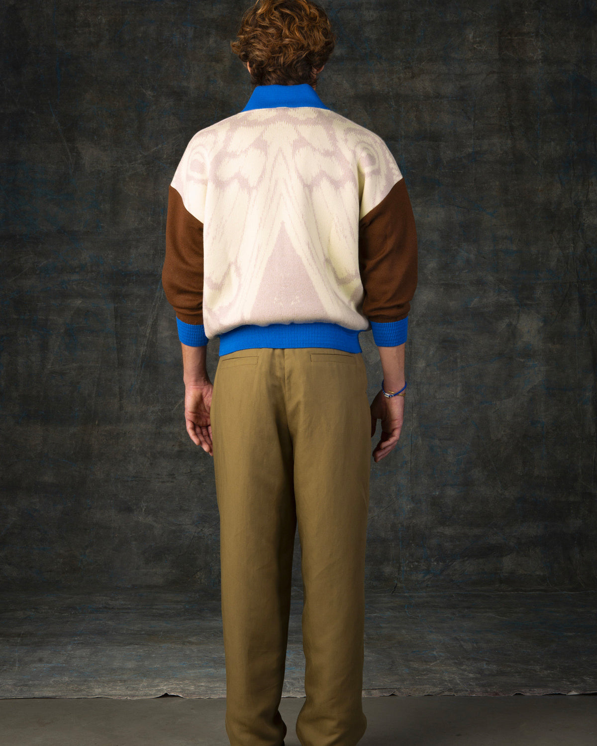 Male model facing away from the camera wearing an ecru and brown bomber jacket with cobalt blue details. There is a subtle organic pattern inspired by a butterfly's wings on the back of the jacket.