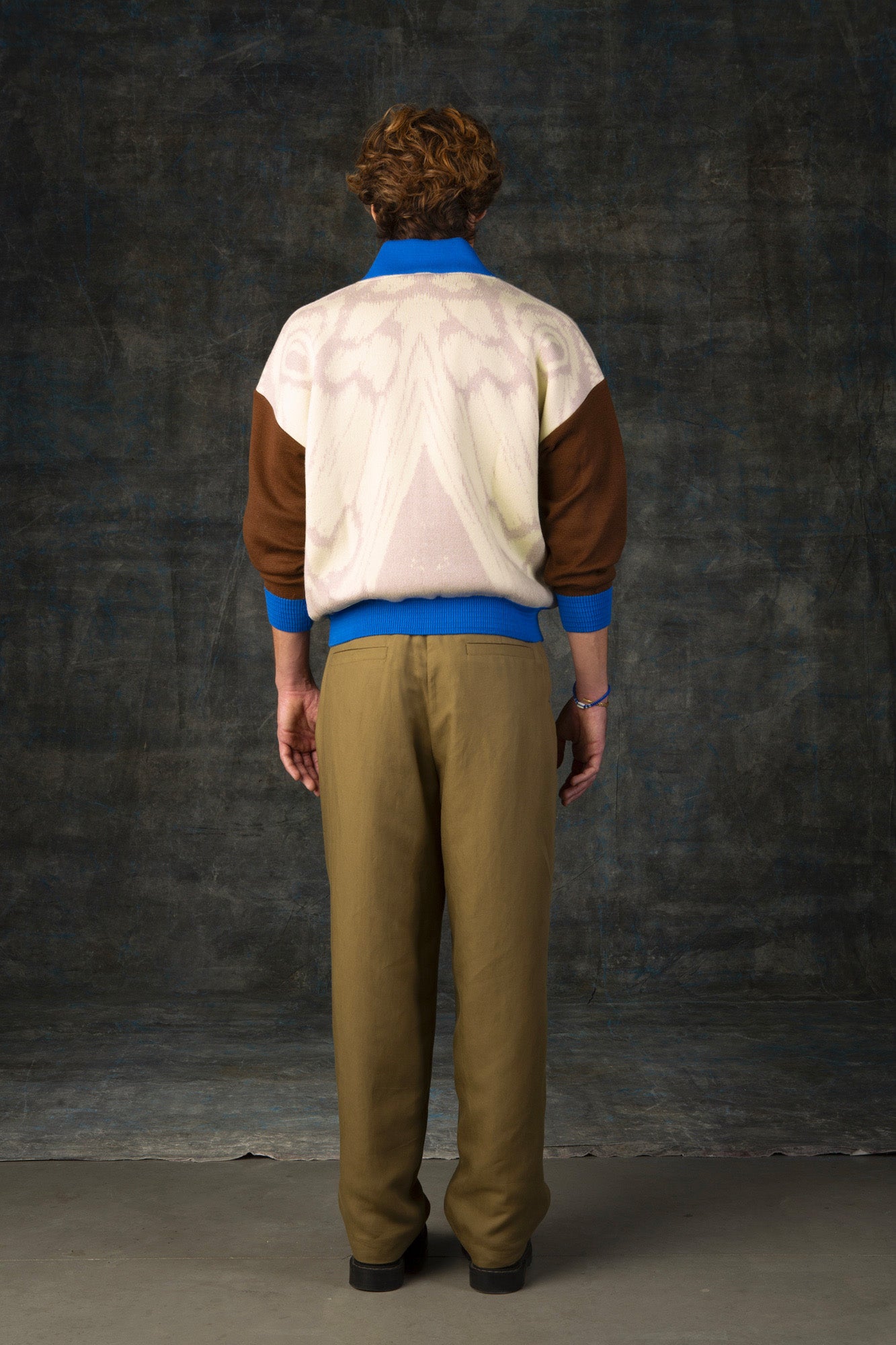 Male model facing away from the camera wearing an ecru and brown bomber jacket with cobalt blue details. There is a subtle organic pattern inspired by a butterfly's wings on the back of the jacket.