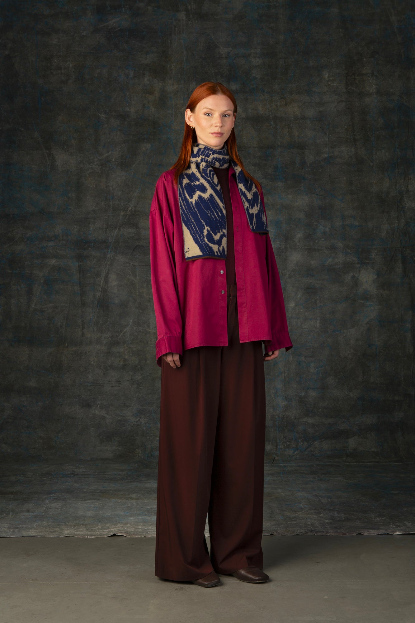 Female model wearing a fuchsia outfit with a dark blue and beige mini scarf draped around her neck, against a dark background.