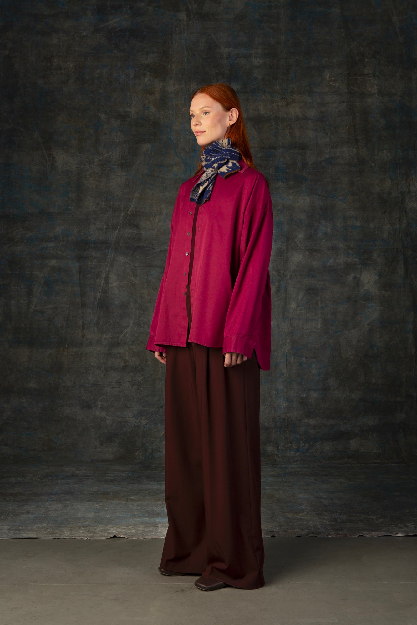 Female model wearing a fuchsia outfit with a dark blue and beige mini scarf closely tied around her neck, against a dark background.