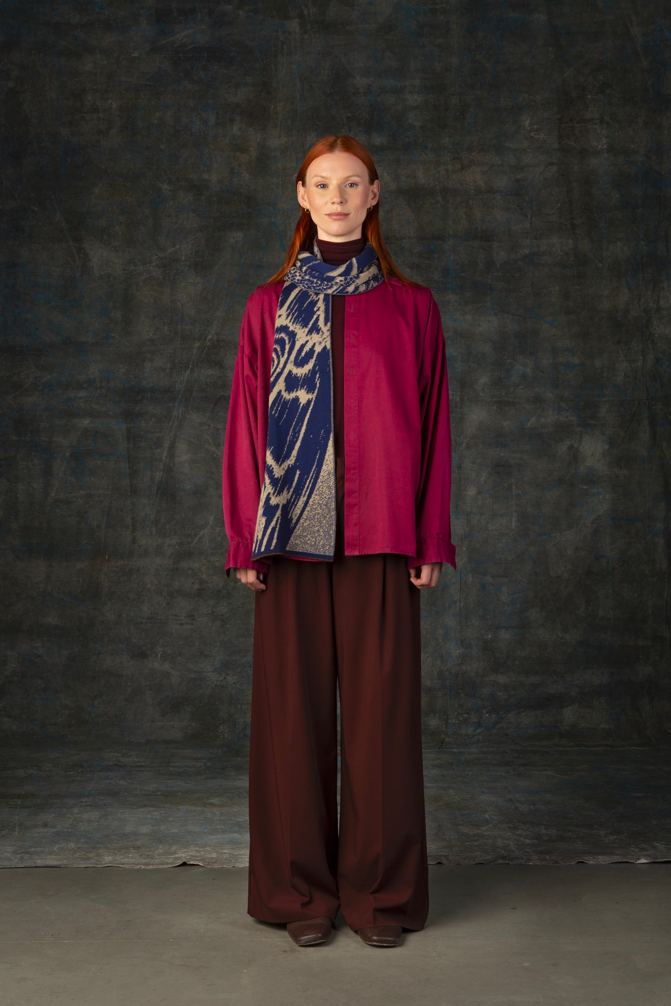 Female model wearing a fuchsia outfit with a dark blue and beige midi scarf draped around her neck, against a dark background.