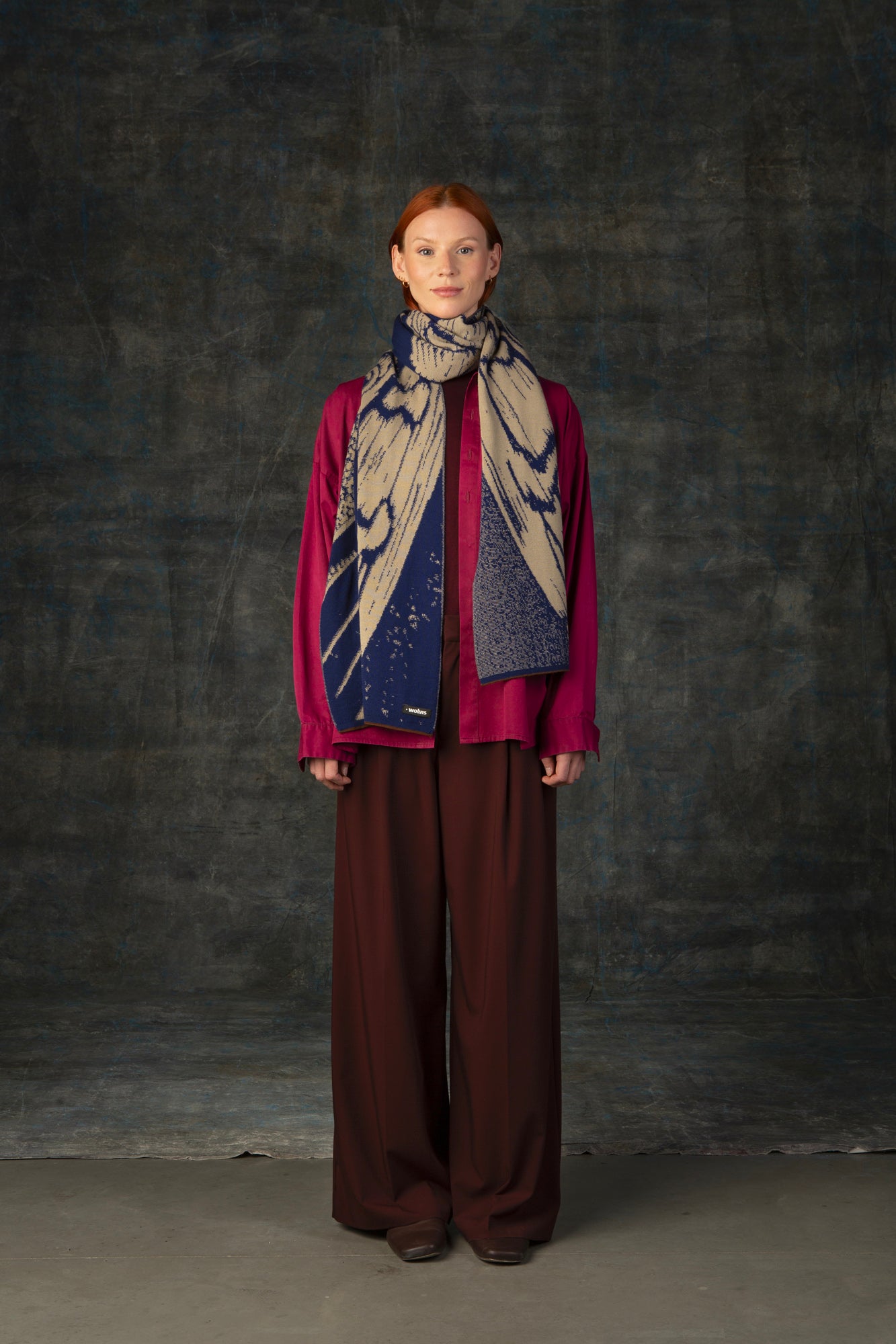 Female model wearing a fuchsia outfit with a dark blue and beige maxi scarf draped around her neck, against a dark background.