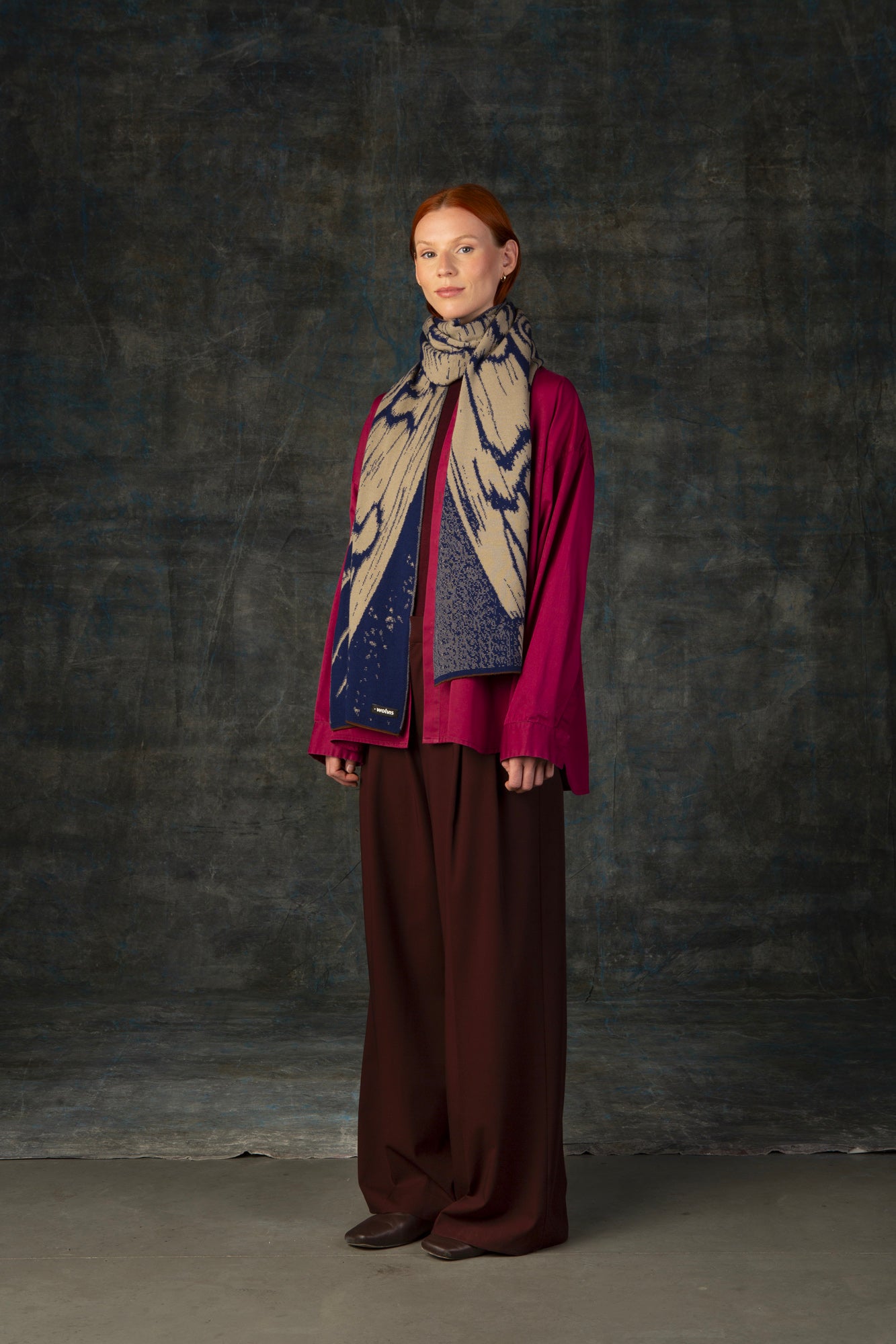 Female model wearing a fuchsia outfit with a dark blue and beige maxi scarf draped around her neck, against a dark background.