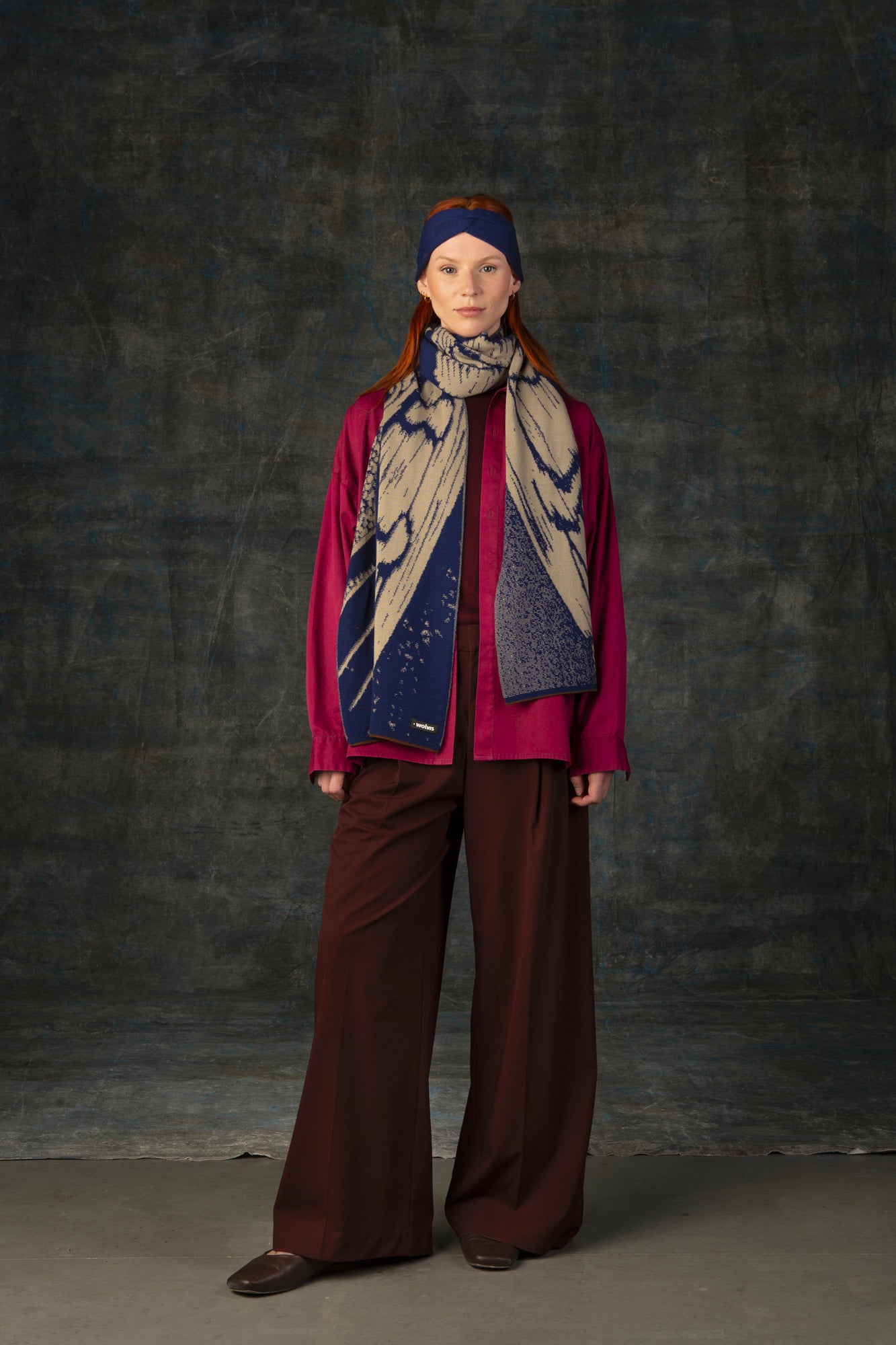 Female model wearing a fuchsia outfit with a dark blue and beige maxi scarf draped around her neck and matching blue headband, against a dark background.