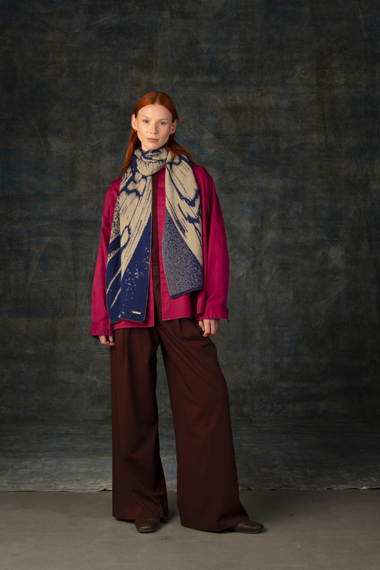 Female model wearing a fuchsia outfit with a dark blue and beige maxi scarf draped around her neck, against a dark background.