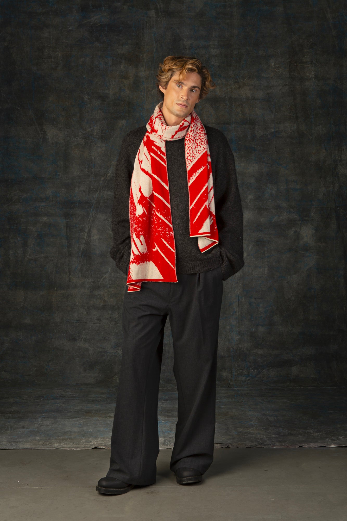 Male model wearing a grey outfit with a bright red and beige maxi scarf with nature-inspired pattern draped around his neck, against a dark background.