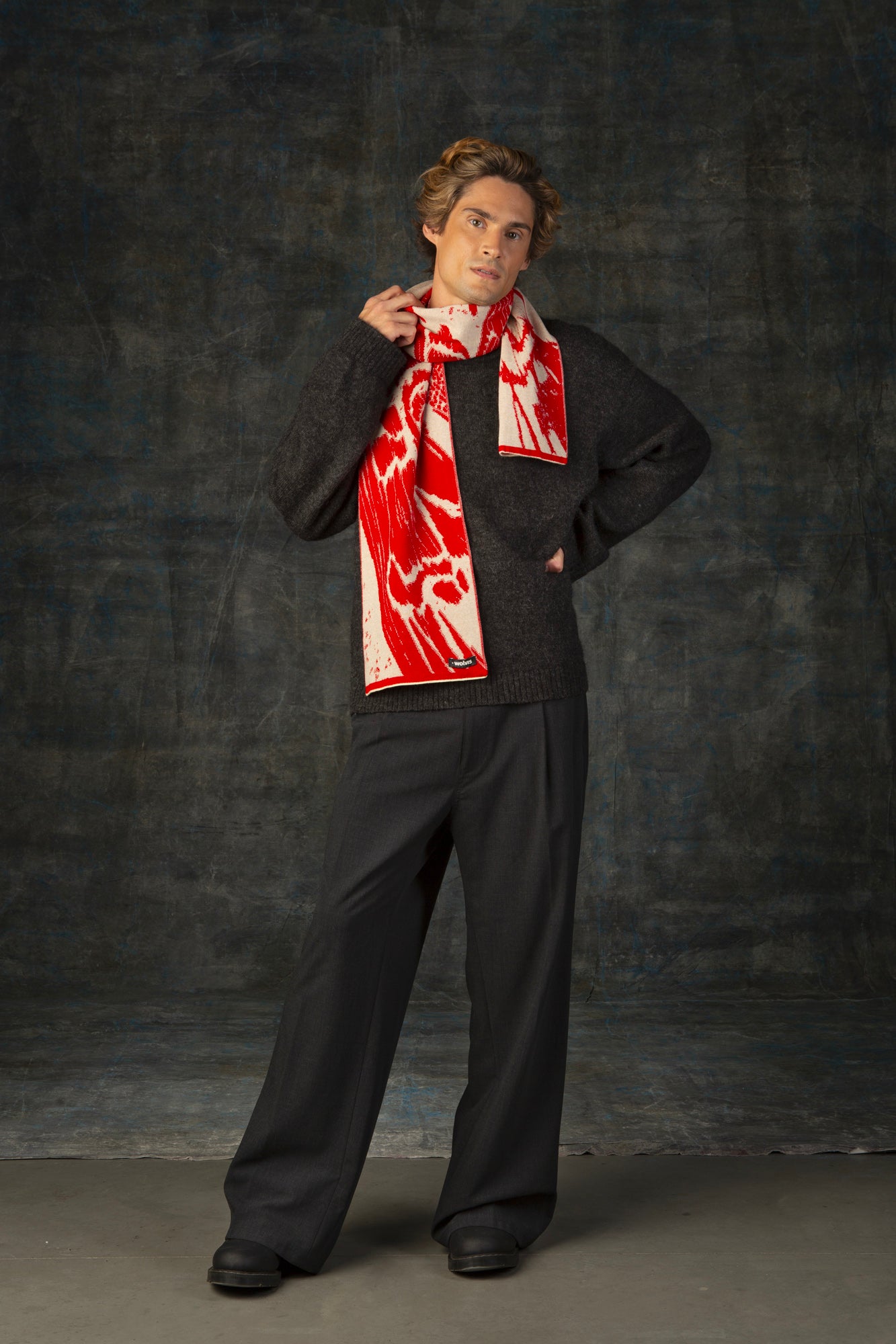 Male model wearing a grey outfit with a bright red and beige midi scarf with nature-inspired pattern draped around his neck, against a dark background.