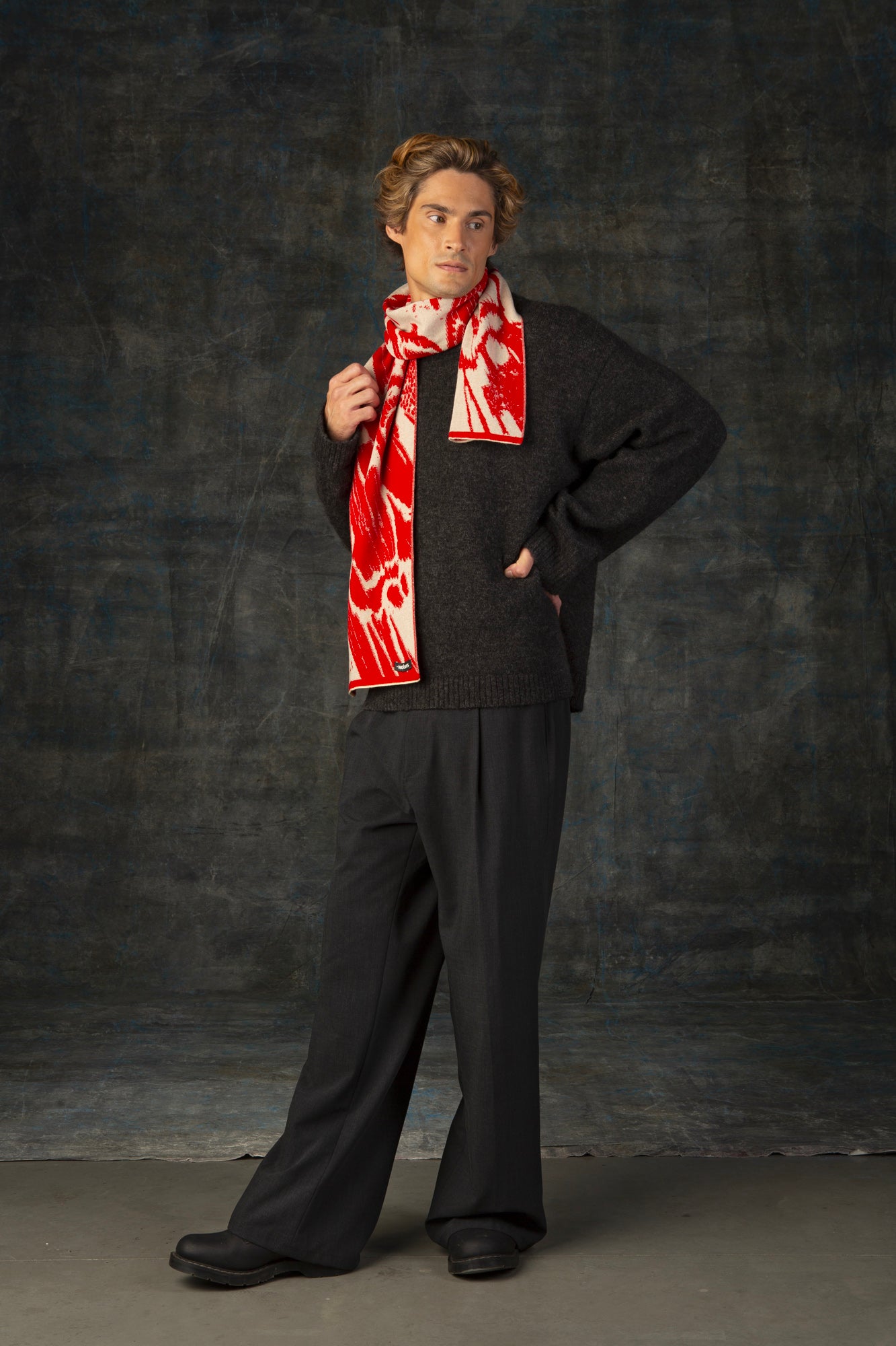 Male model wearing a grey outfit with a bright red and beige maxi scarf with nature-inspired pattern draped around his neck, against a dark background.