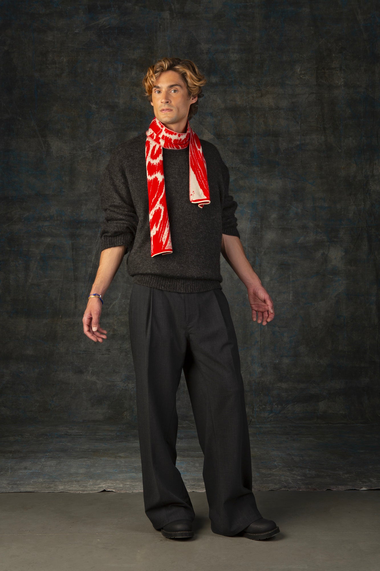 Male model wearing a grey outfit with a bright red and beige mini scarf with nature-inspired pattern draped around his neck, against a dark background.