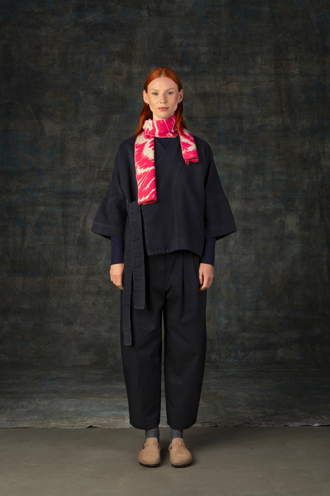 Female model wearing a dark blue outfit with a bright pink and ecru mini scarf, with an organic pattern, draped around her neck.
