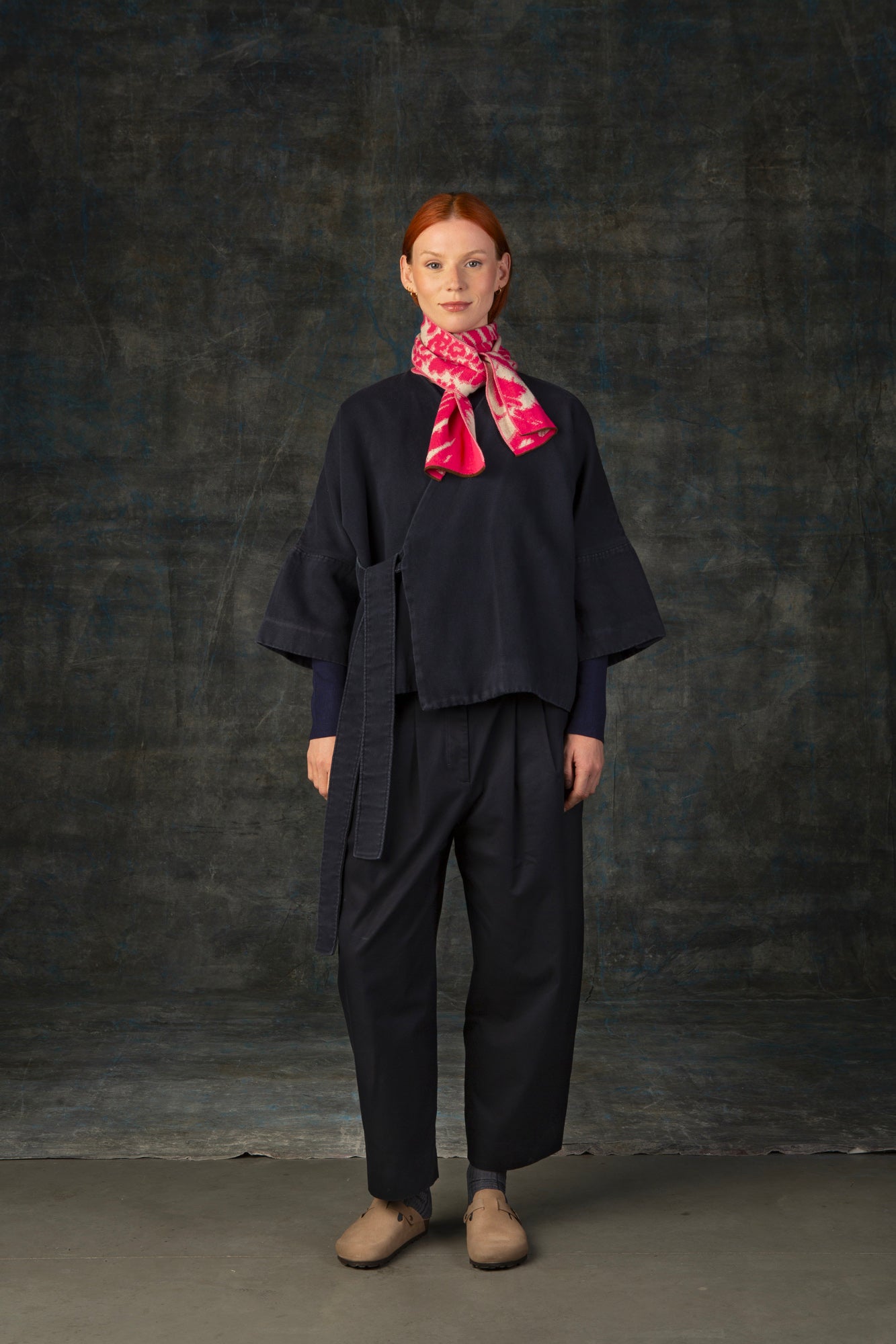 Female model wearing a dark blue outfit with a bright pink and ecru mini scarf, with an organic pattern, tied around her neck.