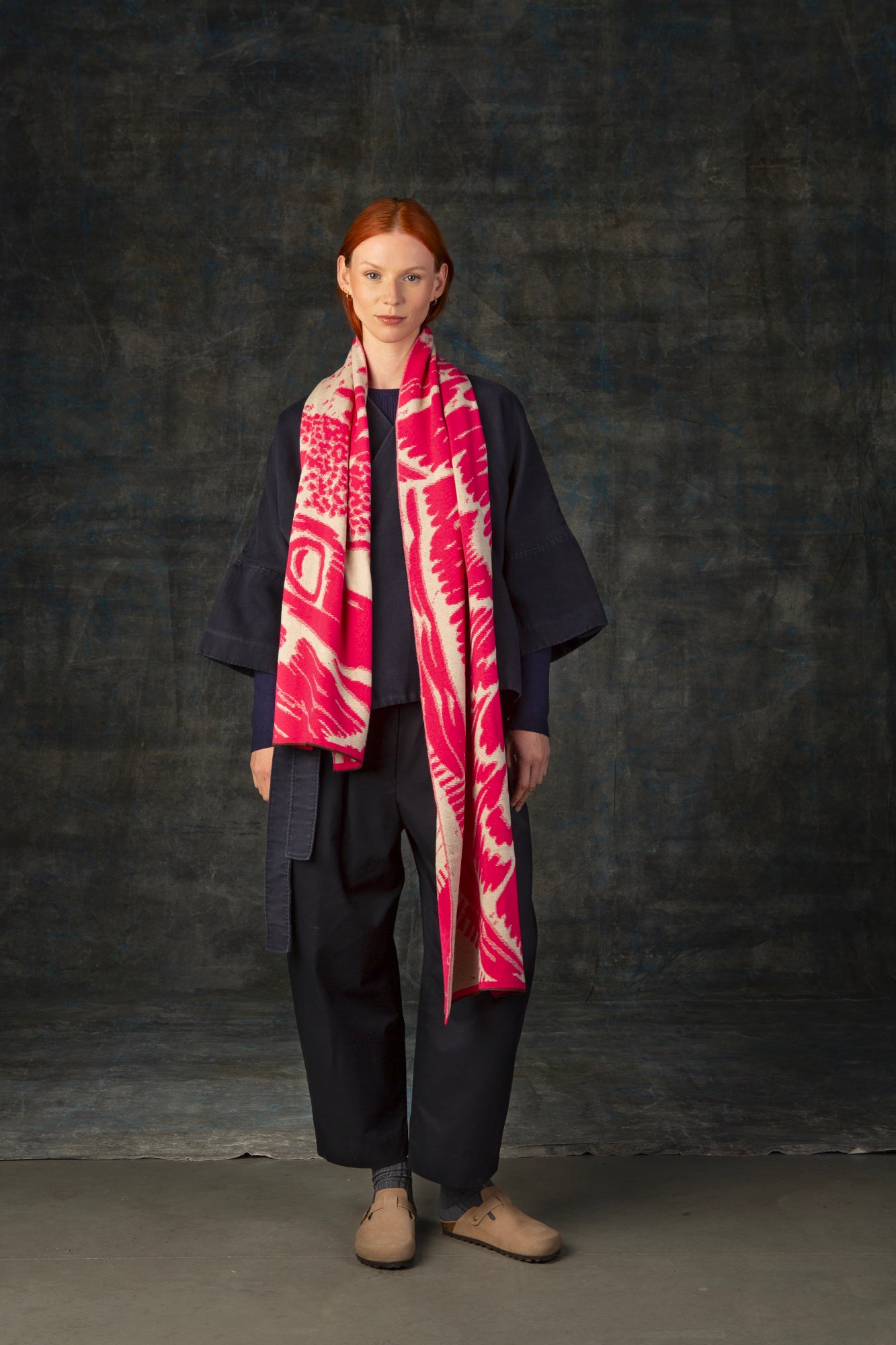 Female model wearing a dark blue outfit with a bright pink and ecru maxi scarf, with an organic pattern, draped around her shoulders.
