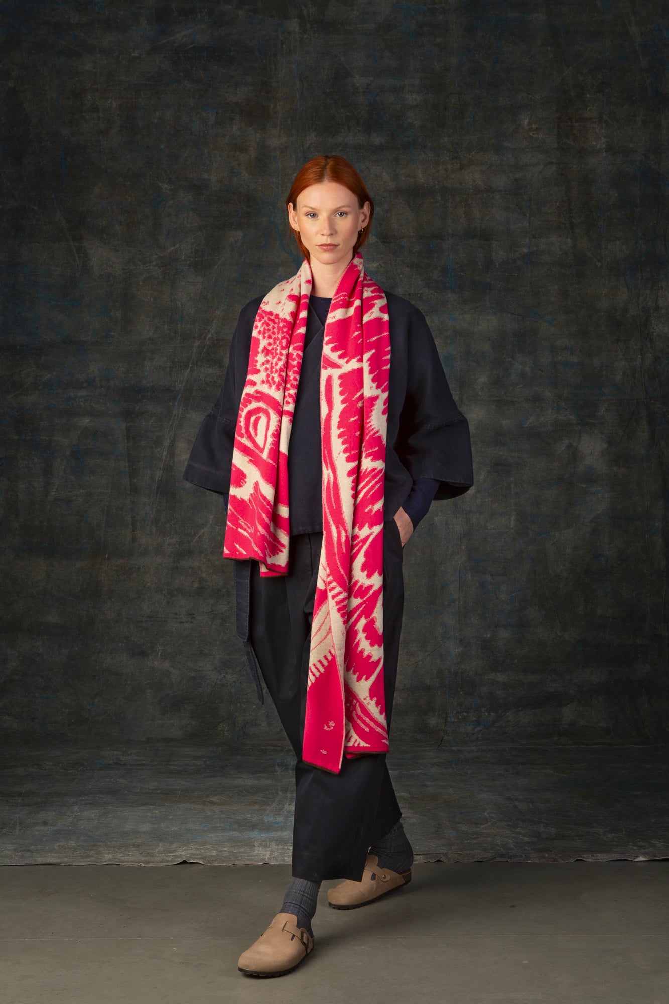 Female model wearing a dark blue outfit with a bright pink and ecru maxi scarf, with an organic pattern, draped around her shoulders.