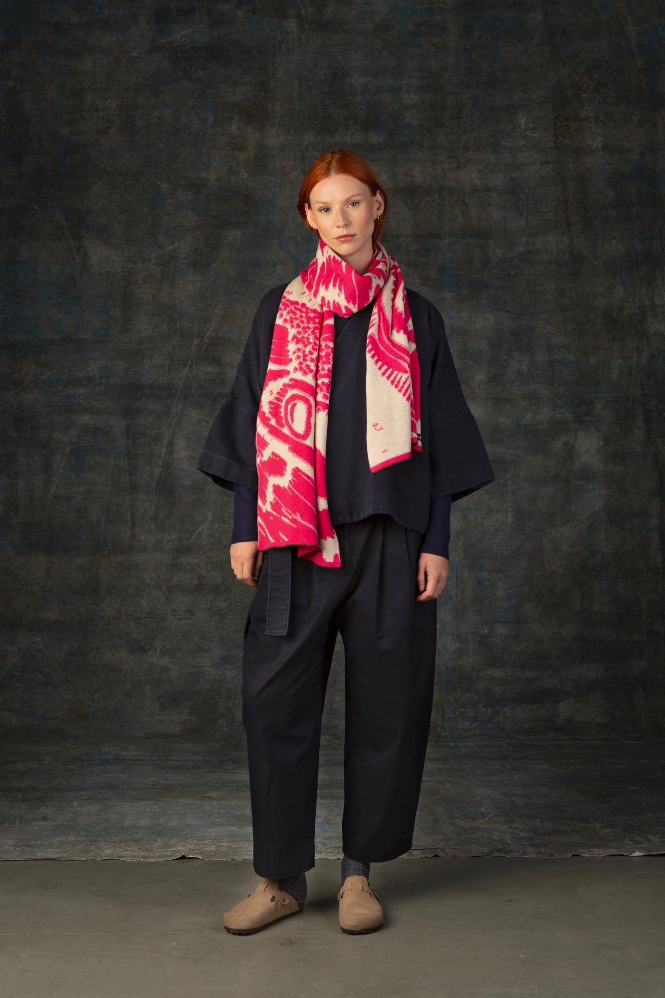 Female model wearing a dark blue outfit with a bright pink and ecru maxi scarf, with an organic pattern, draped around her neck.