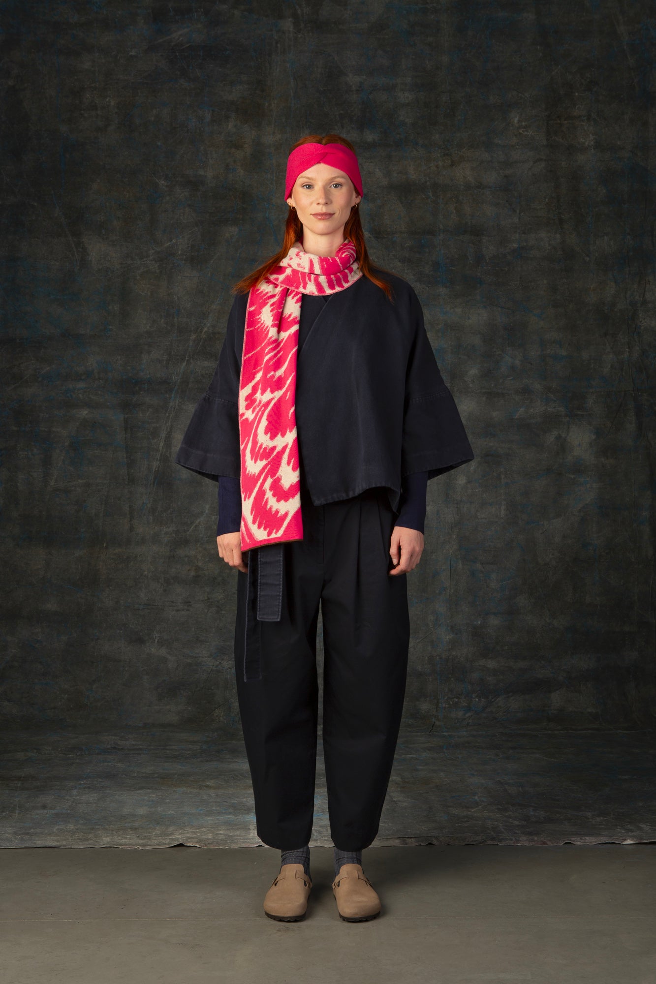 Female model wearing a dark blue outfit with a bright pink and ecru midi scarf, with an organic pattern, draped around her neck and matching bright pink headband.
