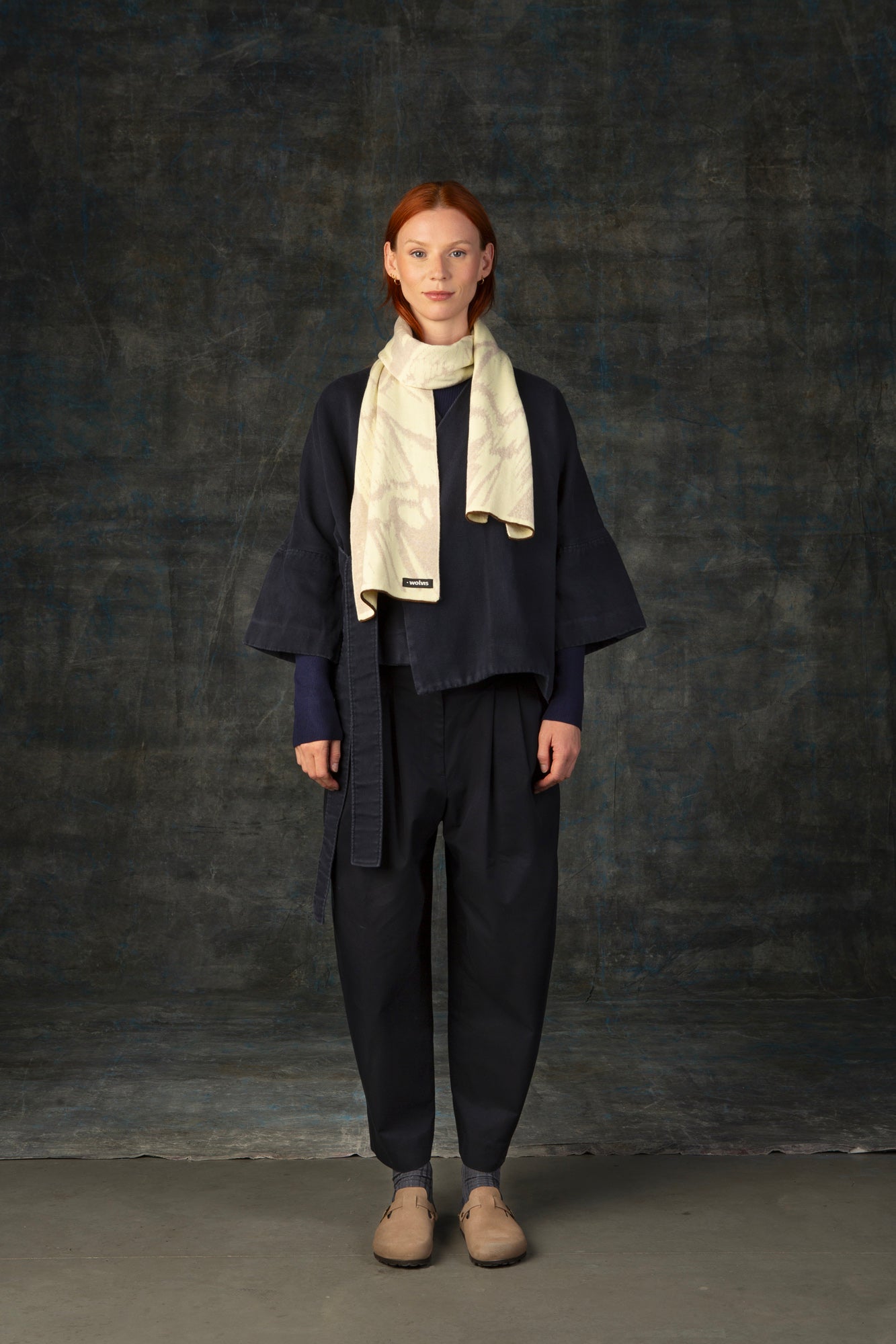 Female model wearing a dark blue outfit with an ecru midi scarf draped around her neck, against a dark background.