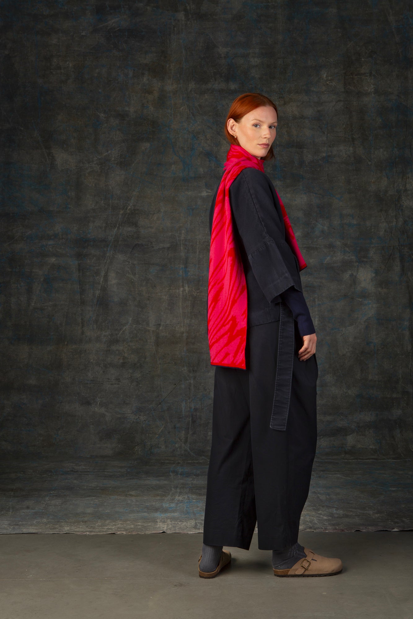 Female model wearing a dark blue outfit with a bright red and pink midi scarf, with nature-inspired pattern draped over her shoulder, against a dark background.
