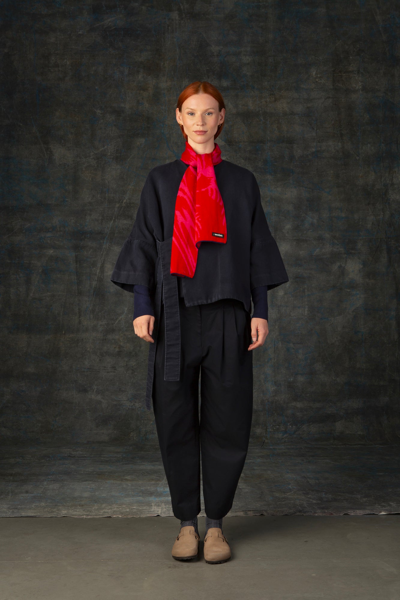 Female model wearing a dark blue outfit with a bright red and pink mini scarf, with nature-inspired pattern loosely tied around her neck, against a dark background.